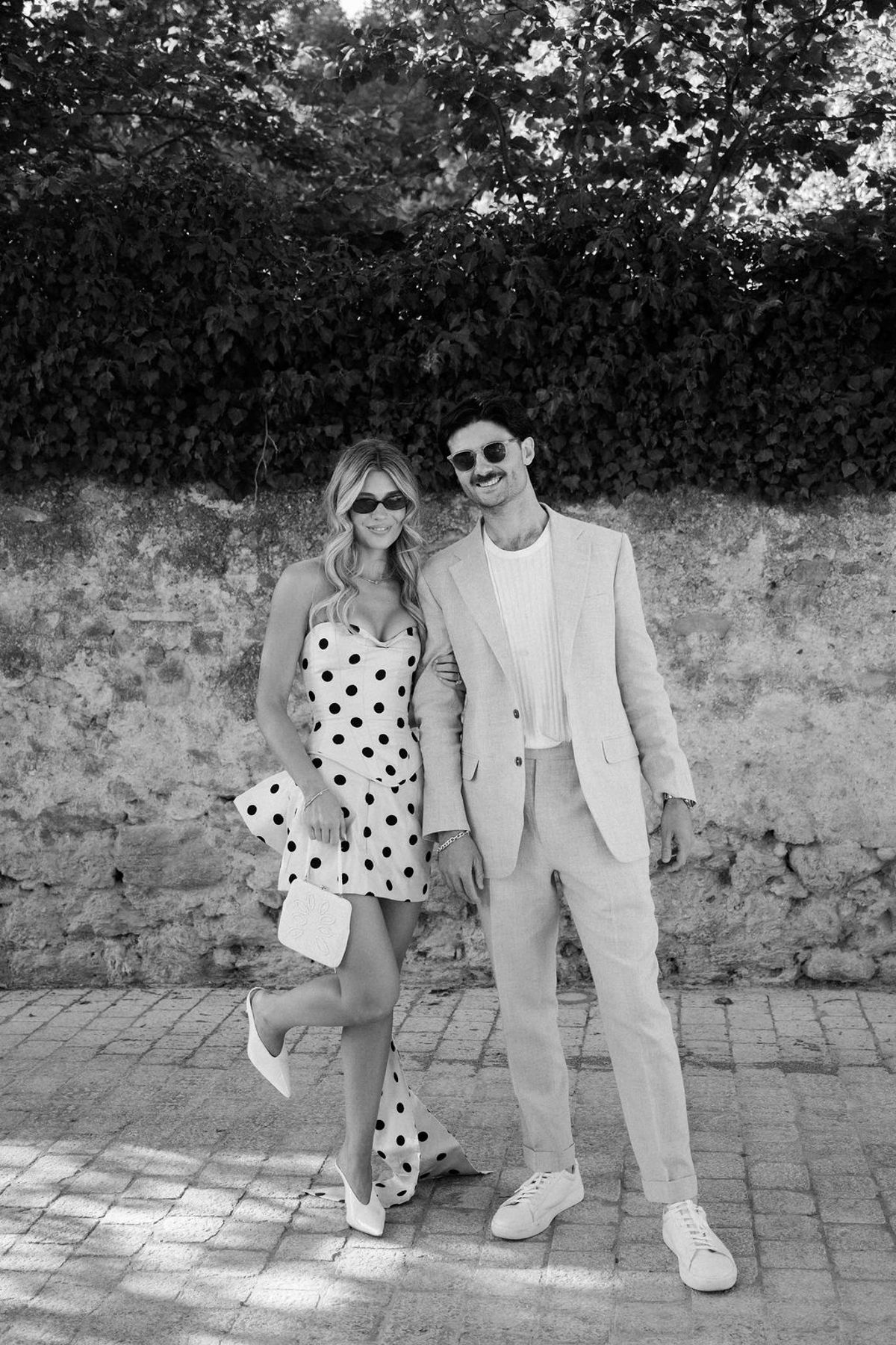 In black and white, a woman in a polka dot dress and a man in a light suit and sunglasses pose side by side.