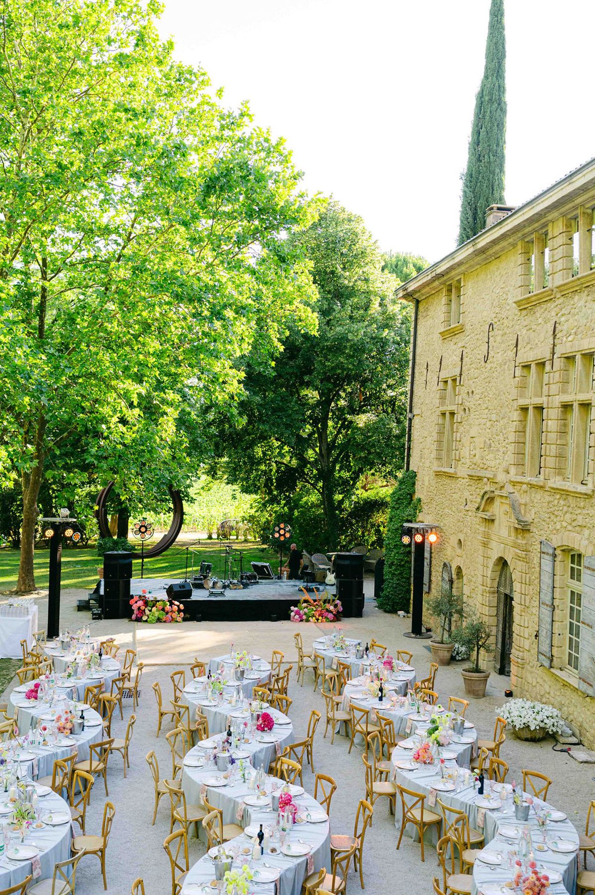 Round tables with pastel flowers and wooden chairs are laid out in a courtyard beside a stone chateau and green trees.