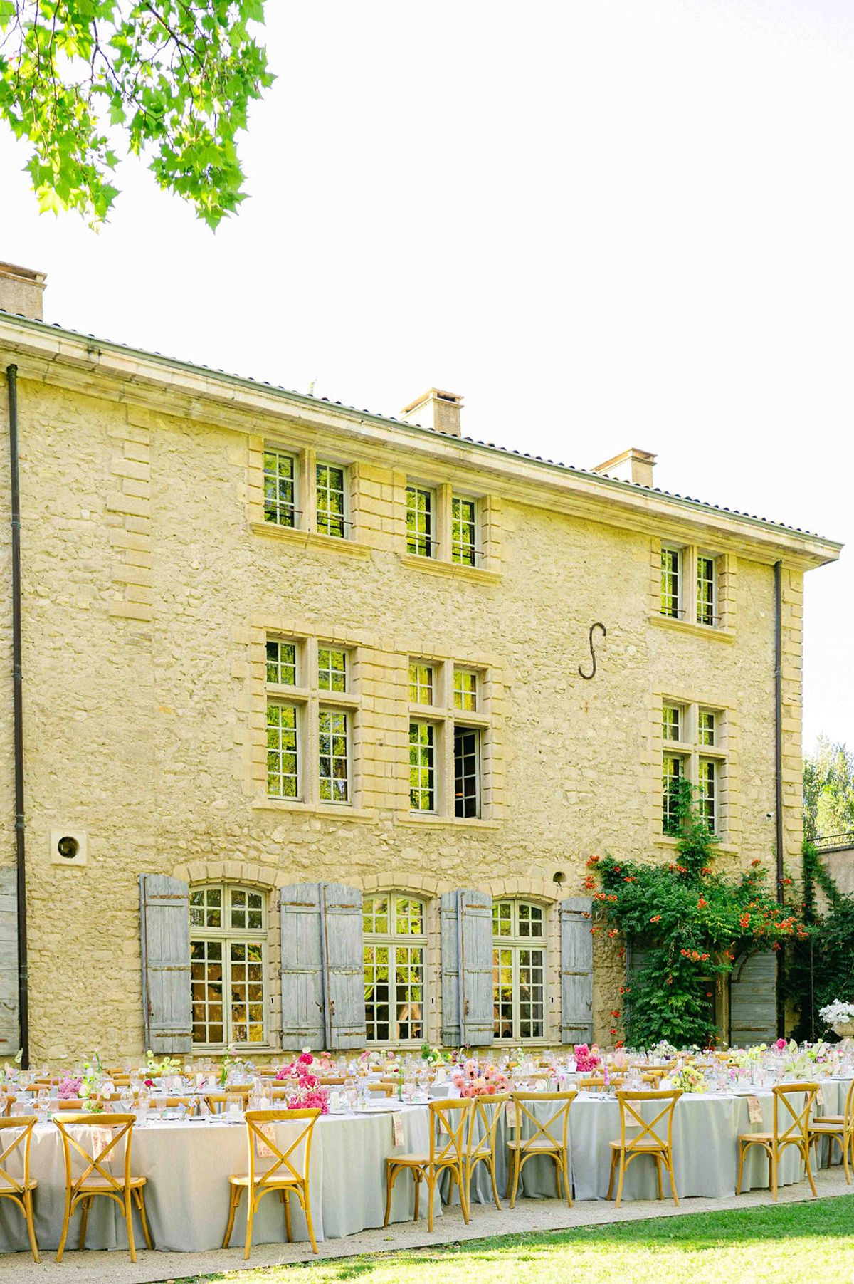 A pale stone building with grey shutters stands behind long dining tables and wooden chairs set out on the grass.