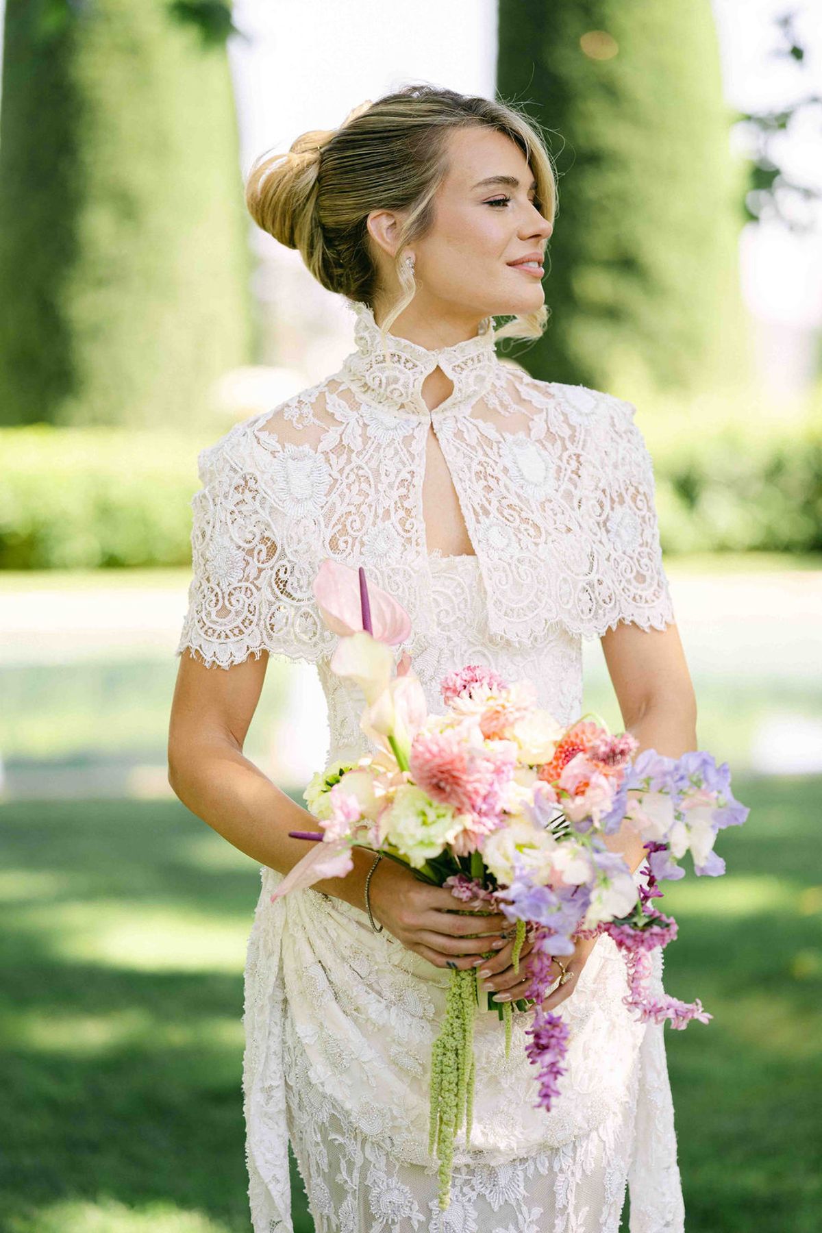 A bride in a lace jacket and gown holds a pastel bouquet, looking away among tall green cypress trees.