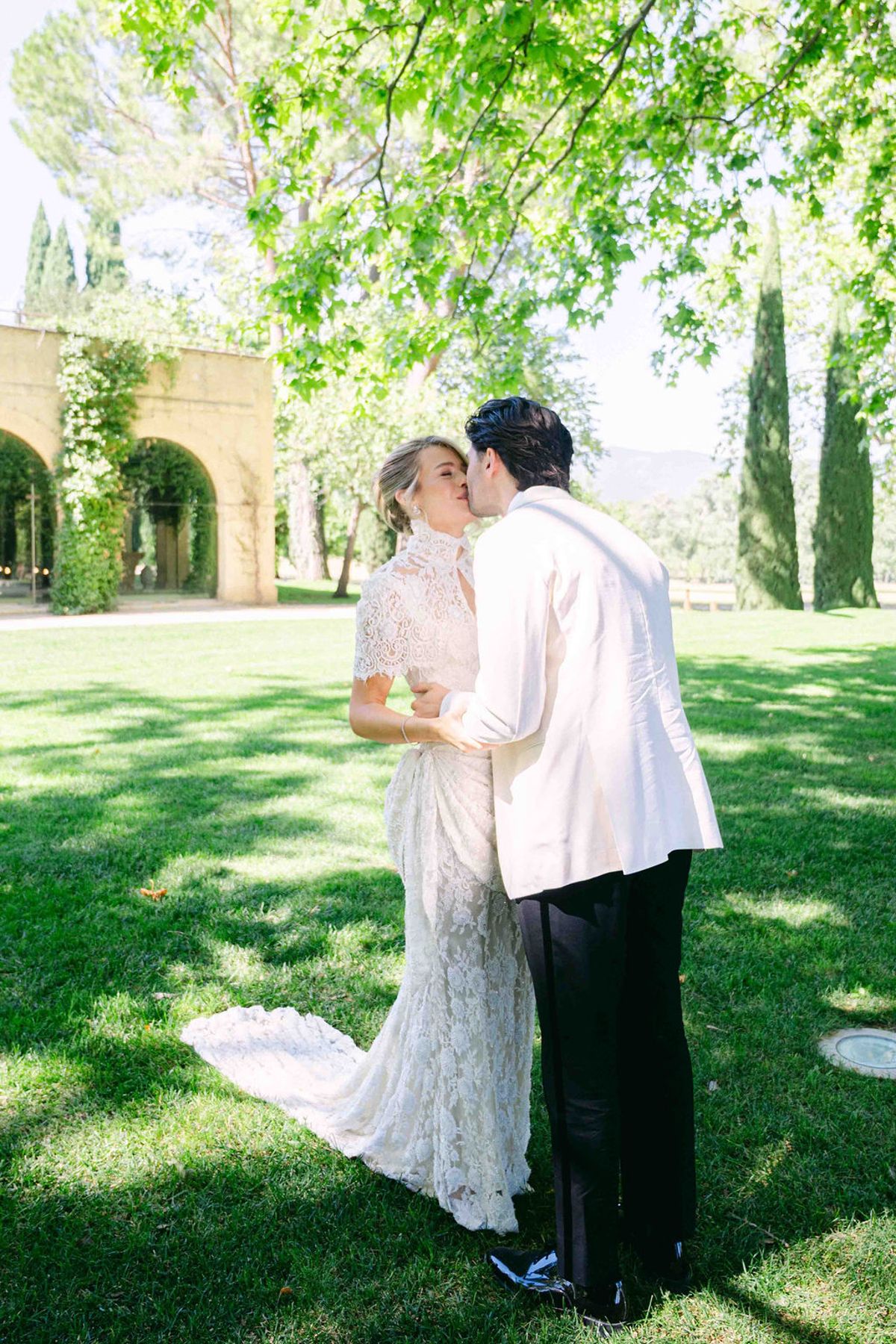 The couple kiss on a sunlit lawn beneath leafy trees, with the bride's long lace train behind her.