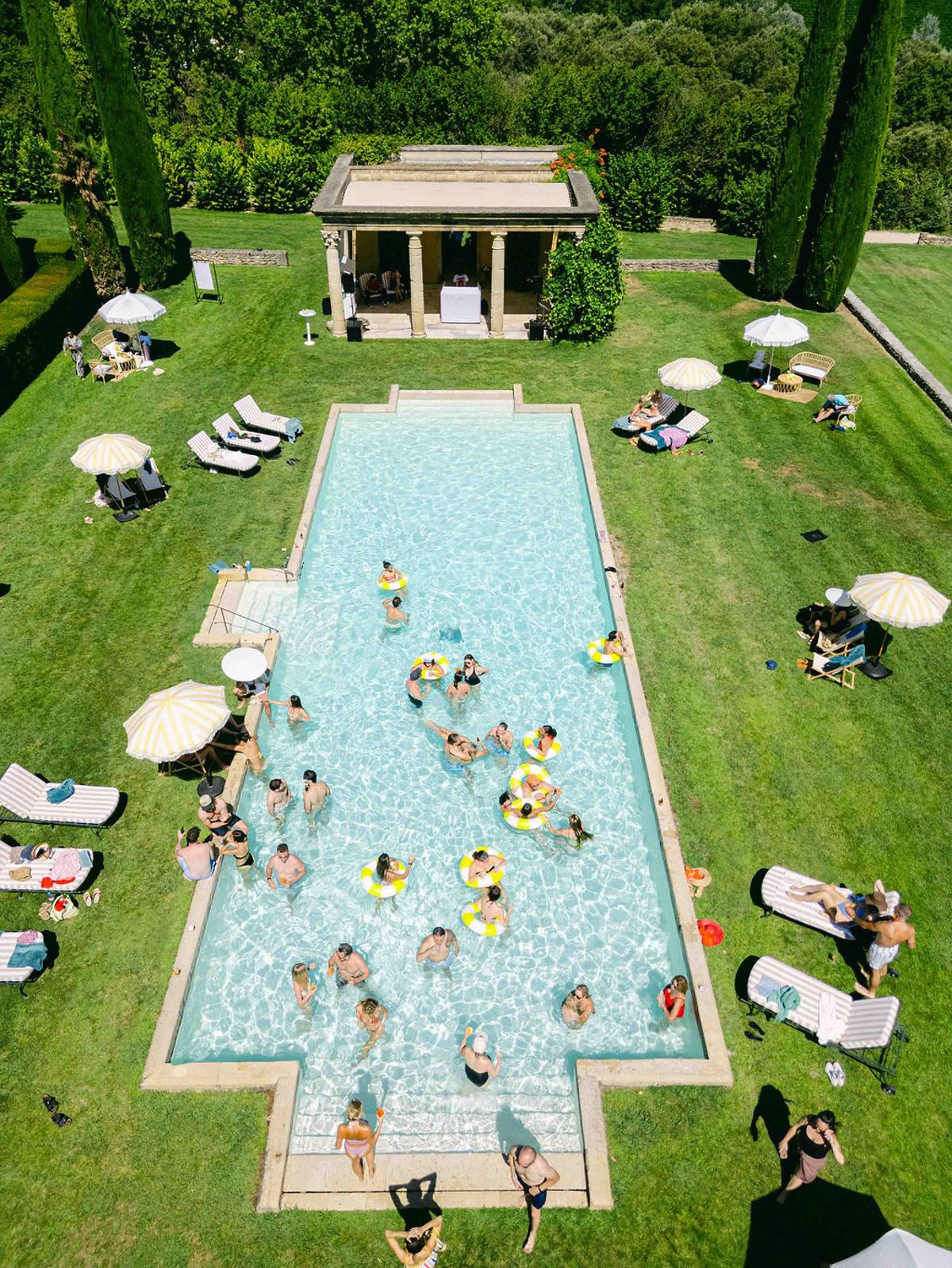 Seen from above, guests swim with colourful floats in a long rectangular pool surrounded by lawn, sunbeds and a small pavilion.