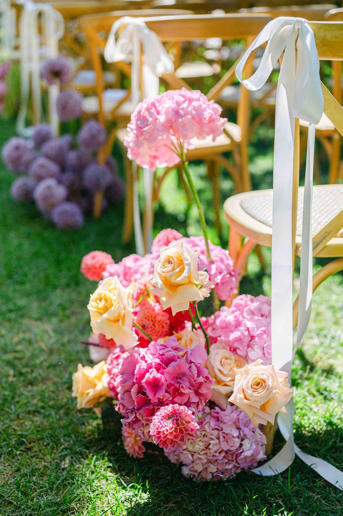A bouquet of pink hydrangeas and roses sits at the end of a wooden chair tied with a long white ribbon.
