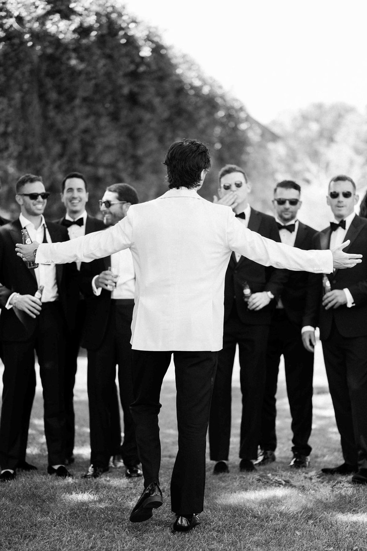 In black and white, the groom in a white jacket, seen from behind, spreads his arms towards groomsmen in dark suits and sunglasses.