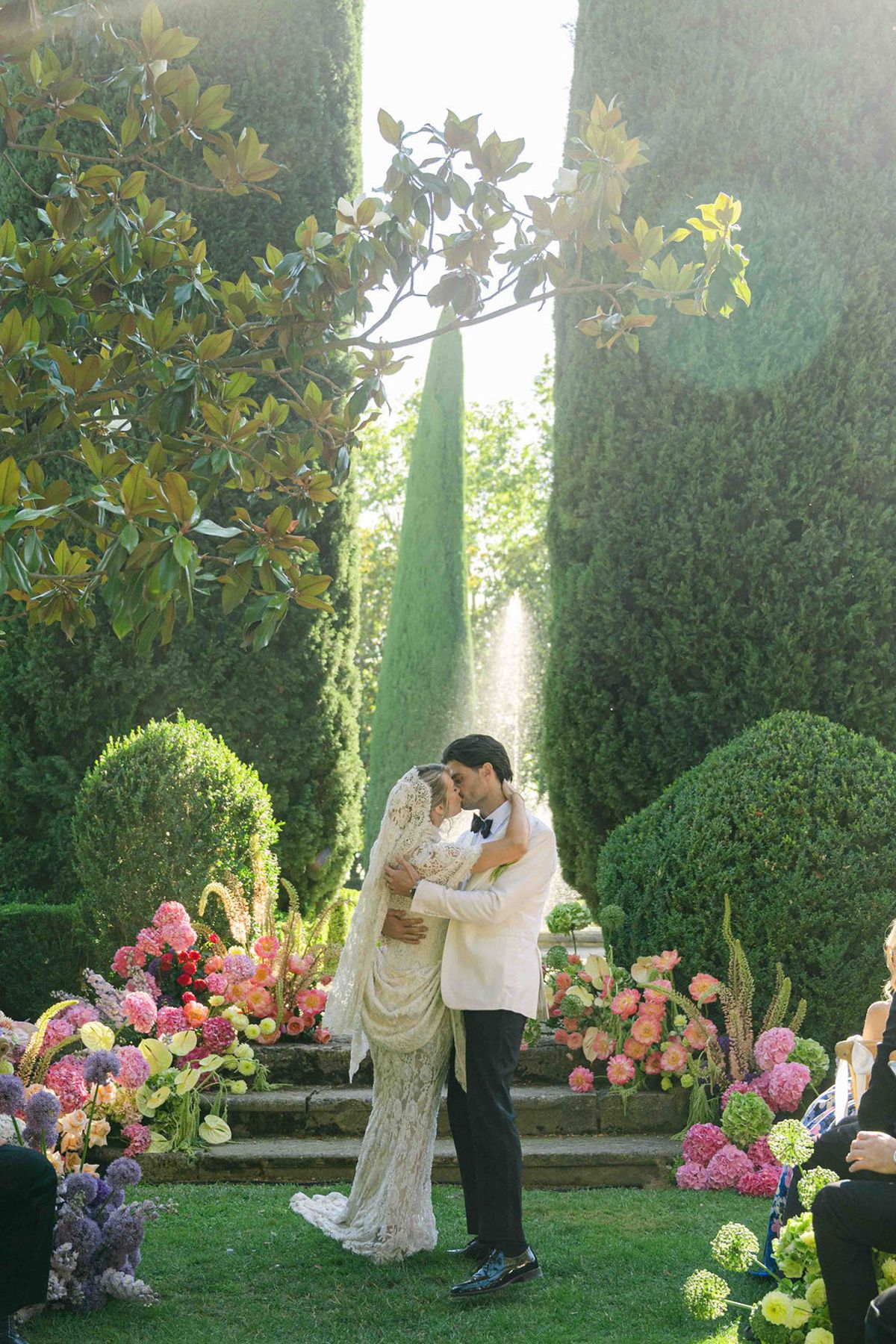 The couple share a kiss in a formal garden between tall hedges, surrounded by bright pink, orange and yellow flower arrangements.