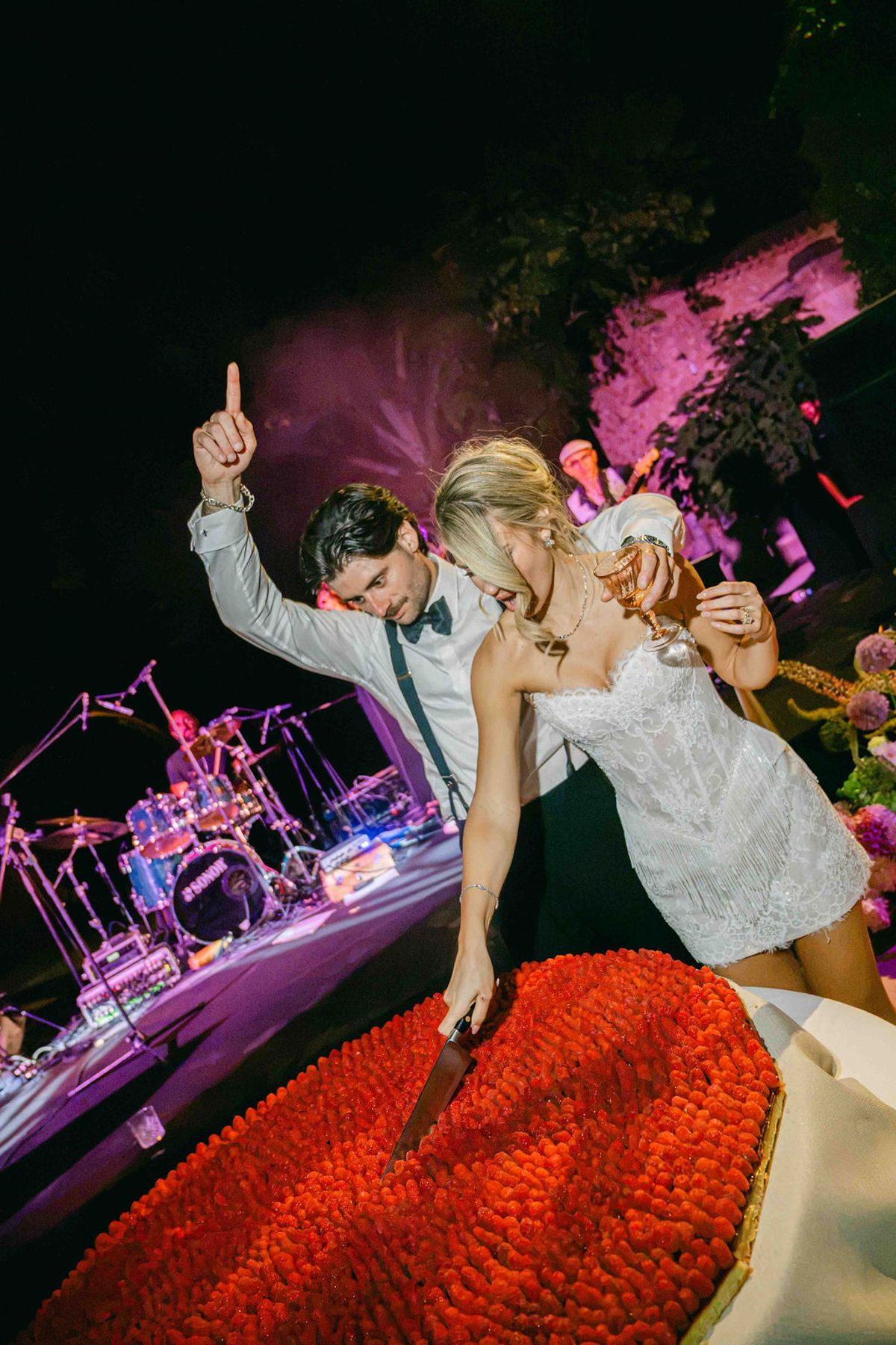 The couple dance beside a large red cake under purple stage lighting, with a drum kit and band equipment behind them.
