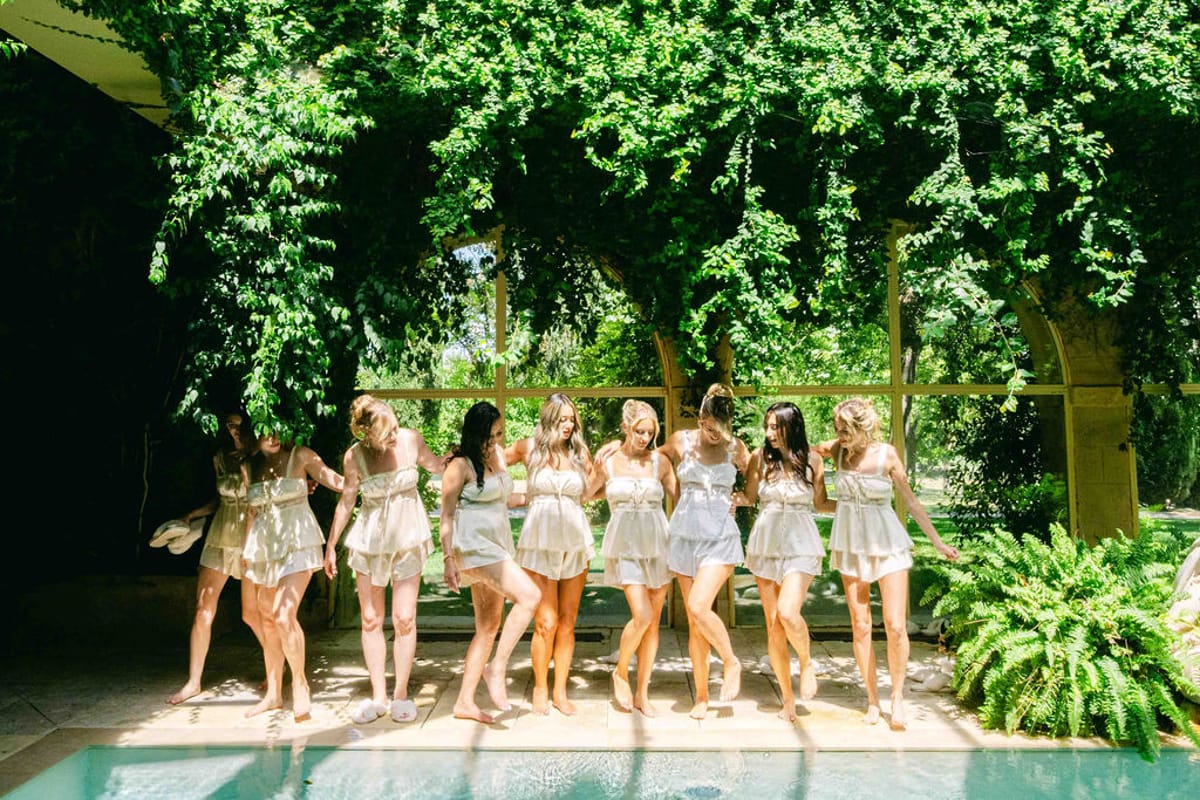 A line of women in matching light pyjama sets walks barefoot beside a pool beneath a canopy of climbing greenery.