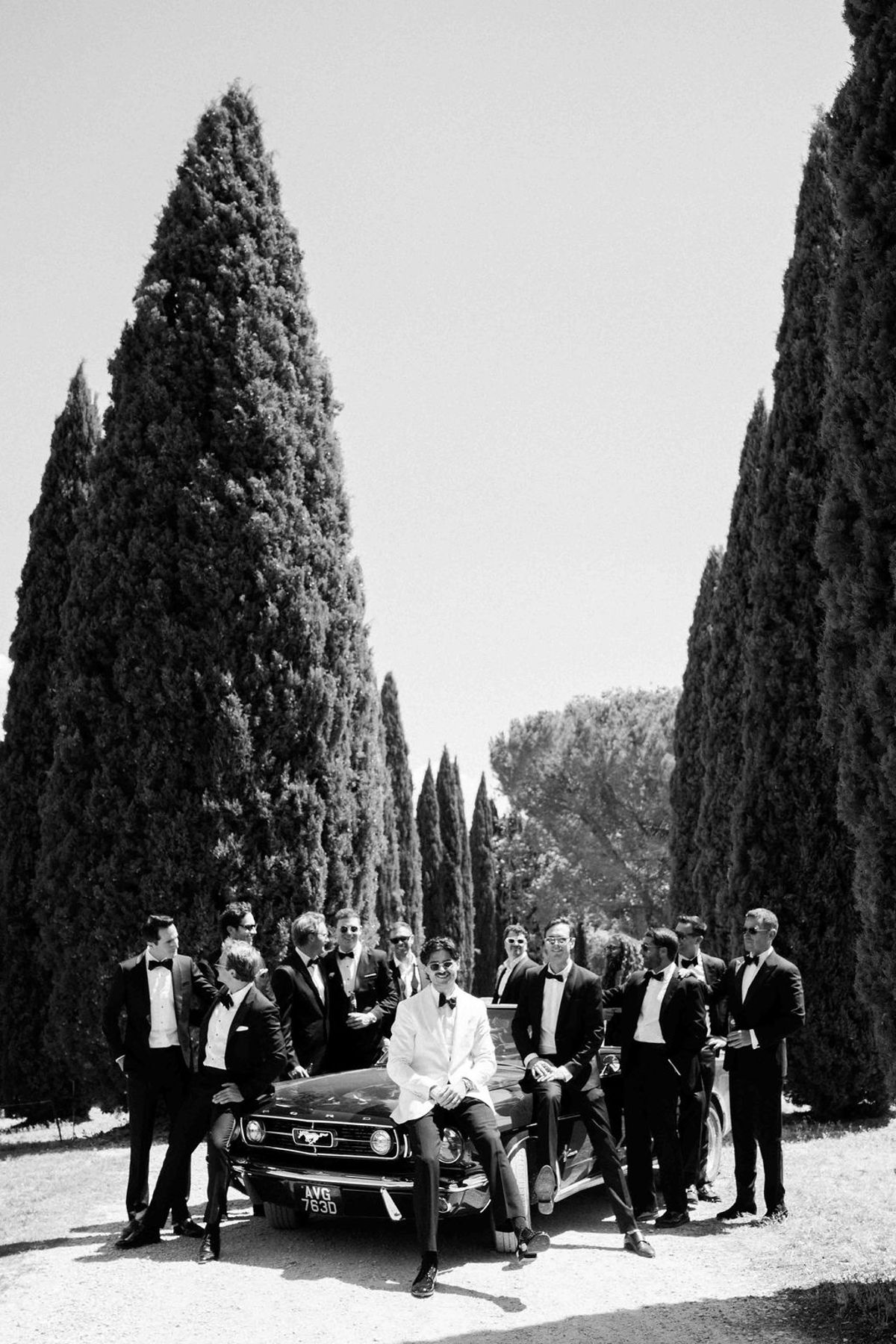 In black and white, men in dark suits stand around a vintage car on a drive lined with tall cypress trees.