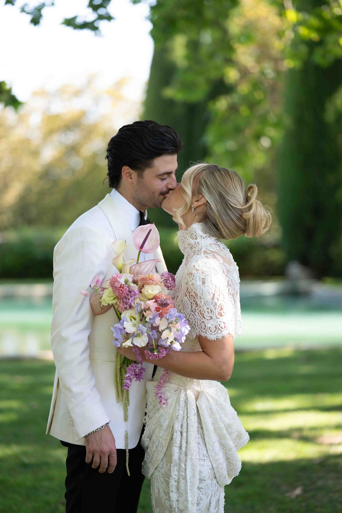 The couple kiss in a green garden, the bride holding a pastel bouquet, with water visible behind them.