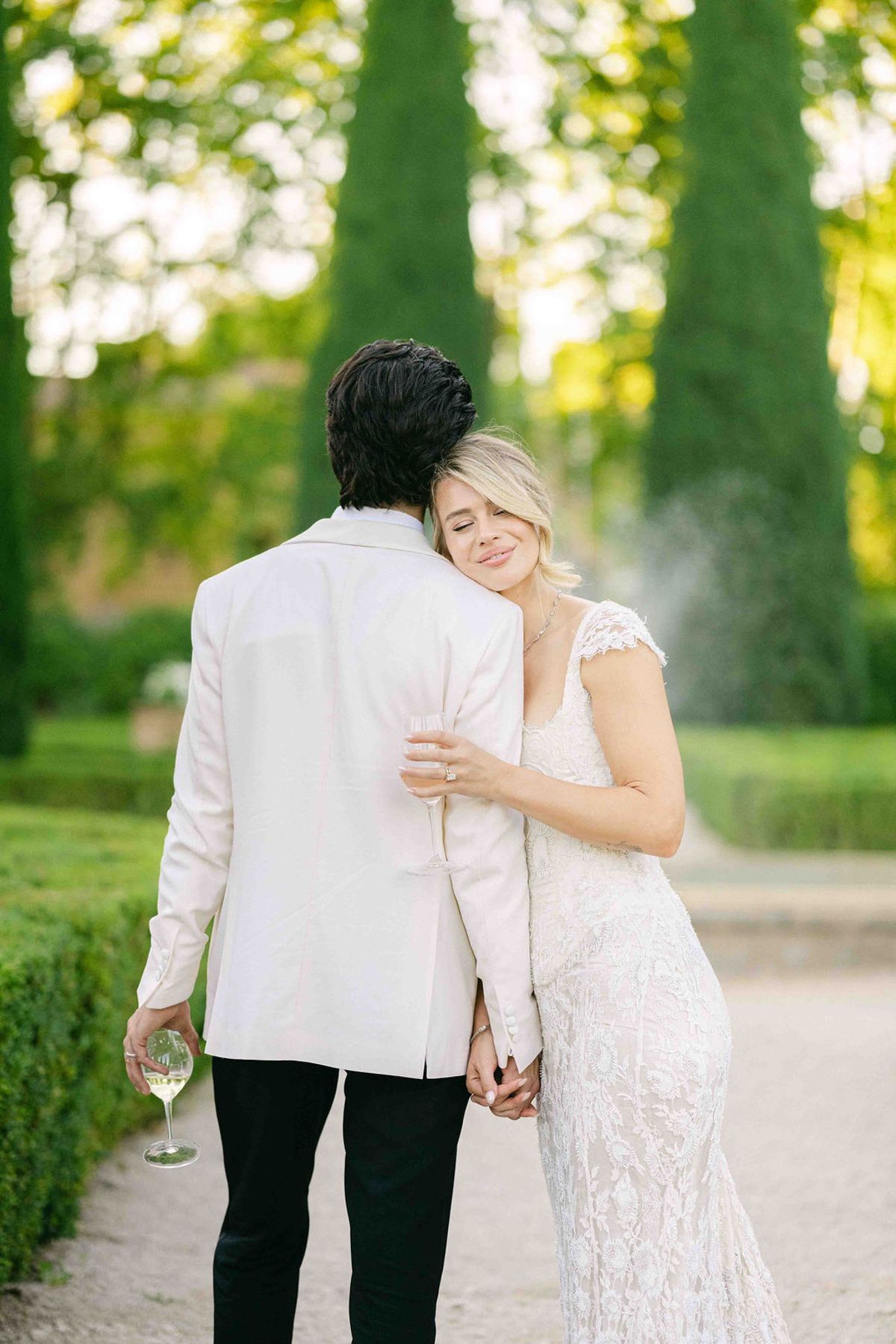 The groom in a white jacket, seen from behind, walks with the bride resting her head on his shoulder in a garden.