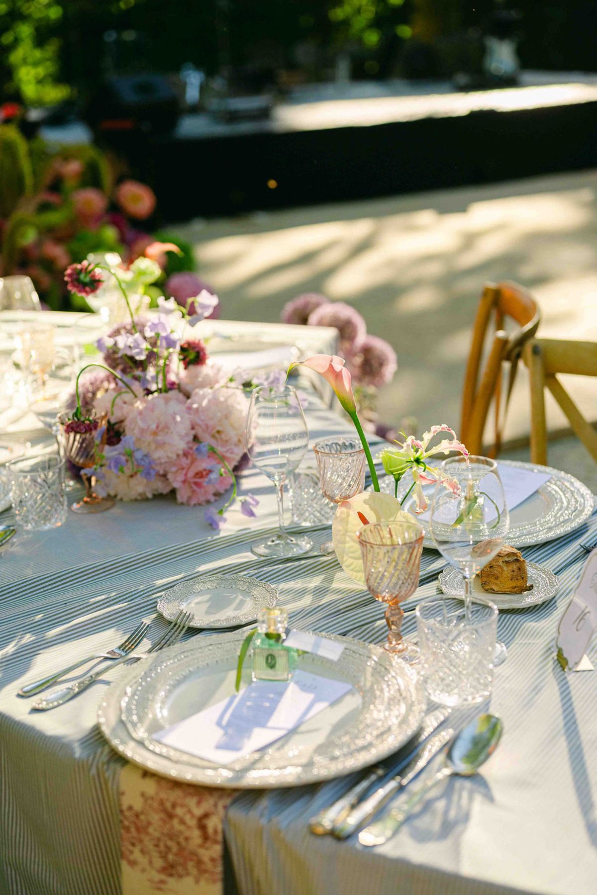 A table with a pale blue cloth holds pastel flowers in small vases, crystal glasses and plates, with wooden chairs beyond.