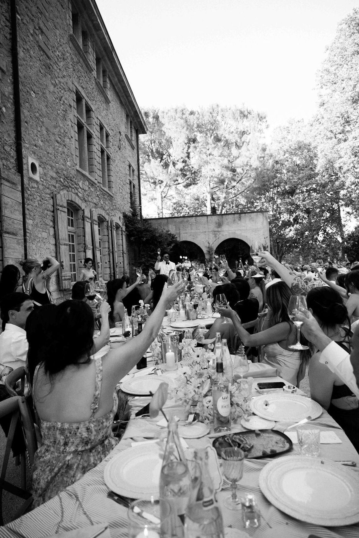 In black and white, guests at a long outdoor dining table raise their arms in a toast beside a stone building.