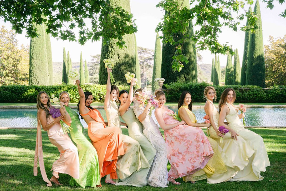 A row of bridesmaids in pastel and coral dresses pose with raised arms on a lawn beside a pool.