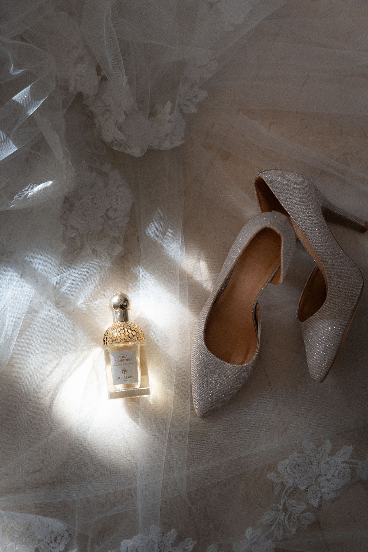 Detail of glitter heels and a perfume bottle resting on lace and tulle in raking light