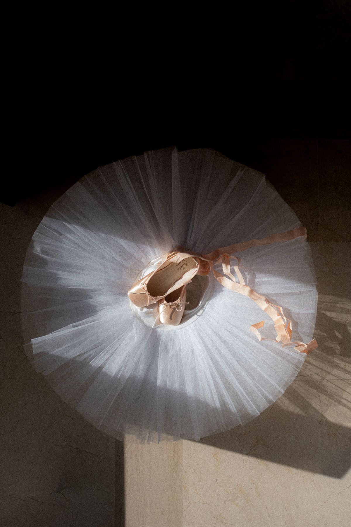 Tulle tutu laid flat with satin pointe shoes at its centre, lit against a dark ground