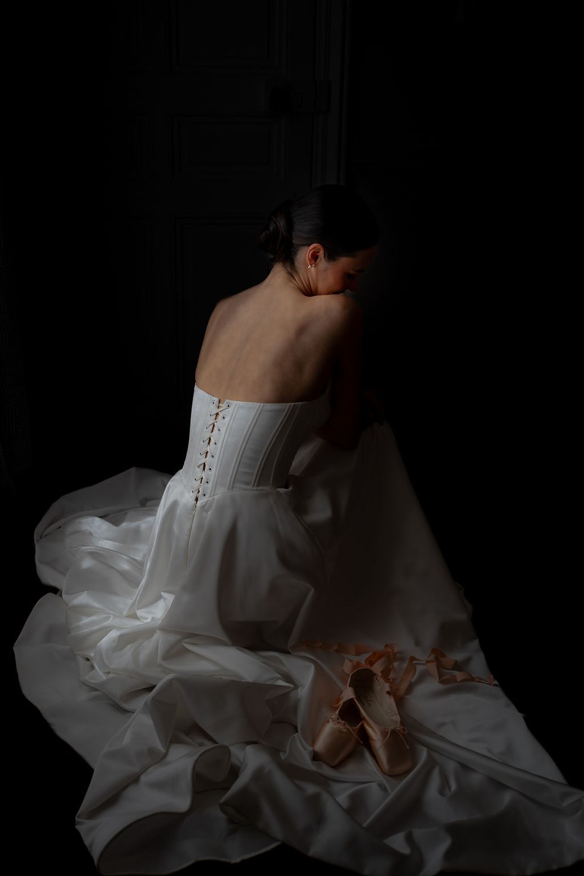 Dancer seated in a corseted gown against a dark backdrop, pointe shoes on the floor beside her
