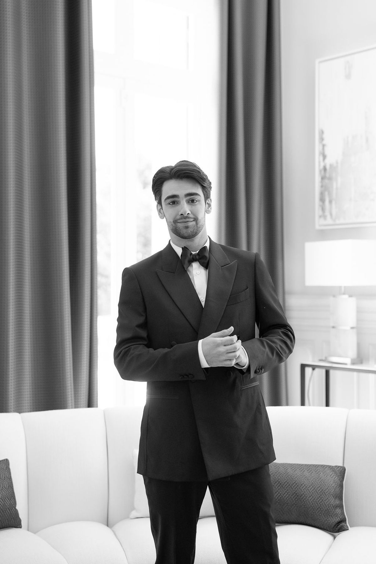 Dancer in black tie standing at a tall curtained window in a bright salon