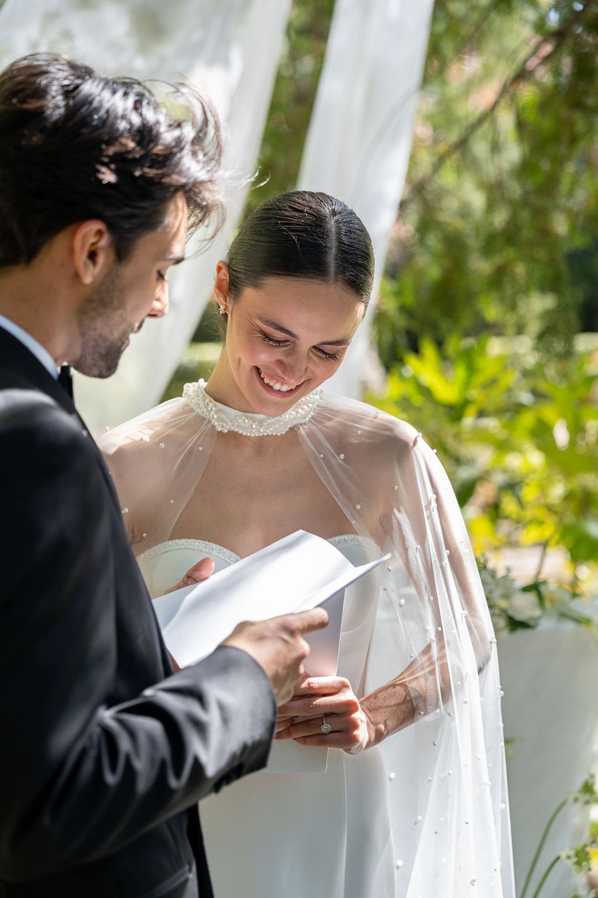 Two dancers reading from a folded card beneath greenery at an outdoor symbolic ceremony