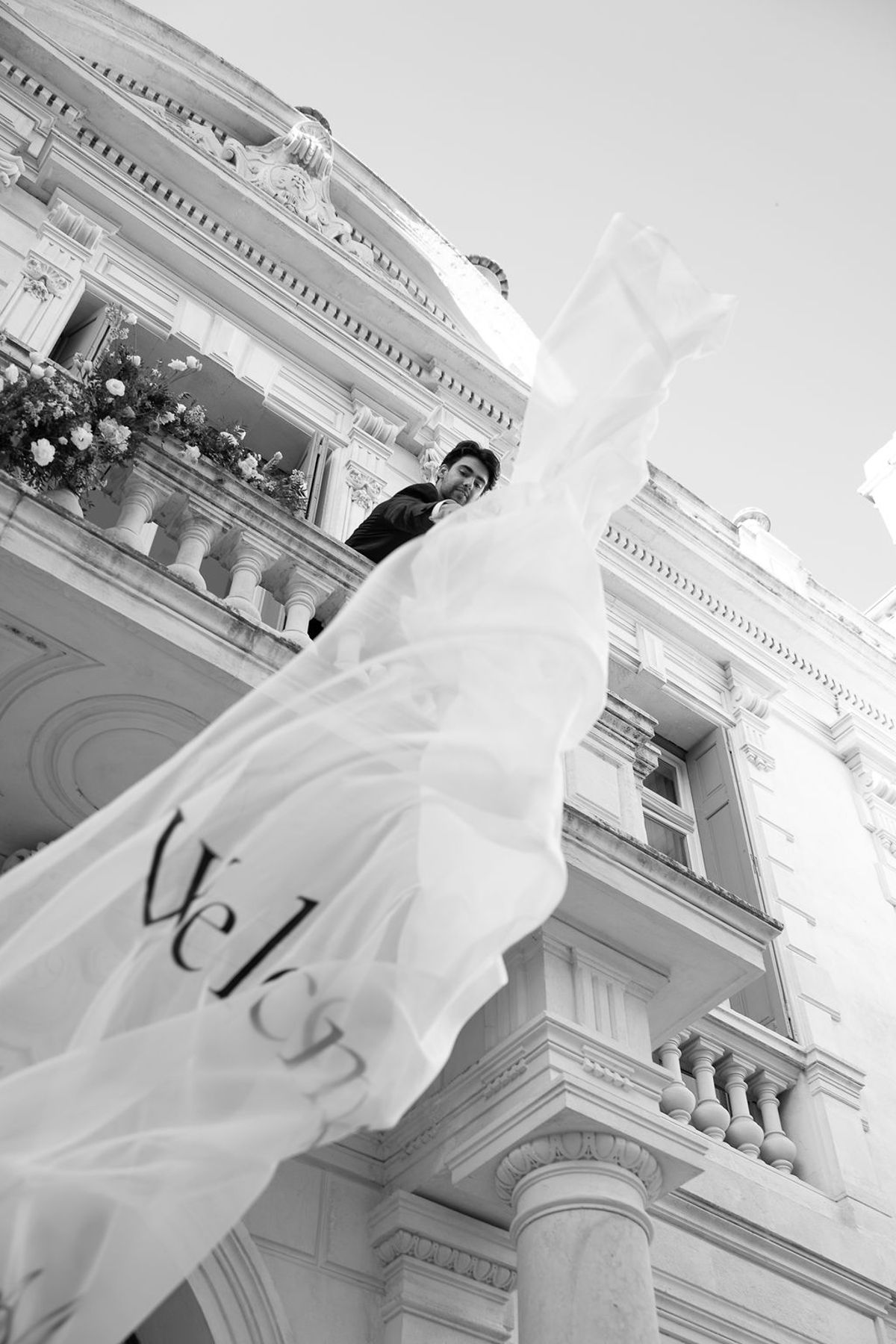 Black and white frame of an embroidered veil billowing from a first-floor balcony at Château des Trois Fontaines, released by a dancer above