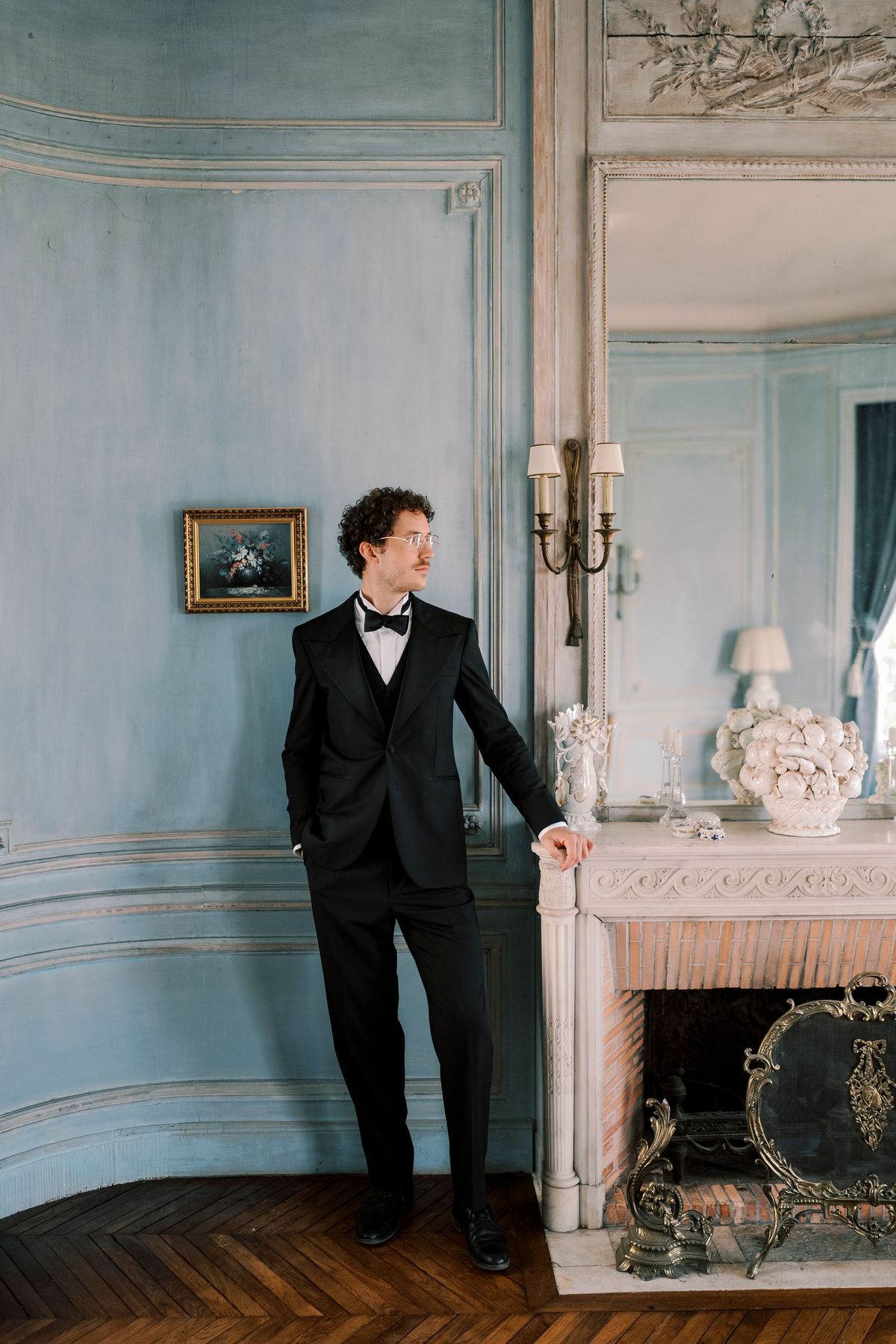 Lucien in black tie standing by the fireplace of a pale blue panelled room
