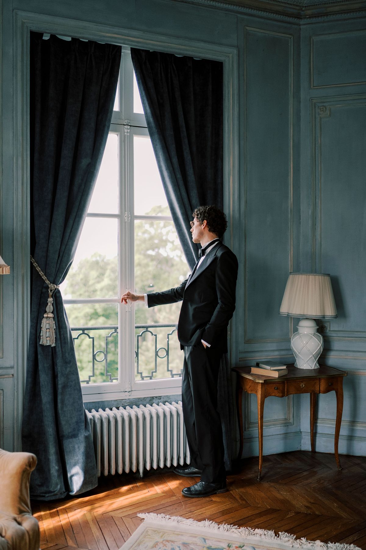 Lucien in black tie drawing back a dark curtain at a tall window