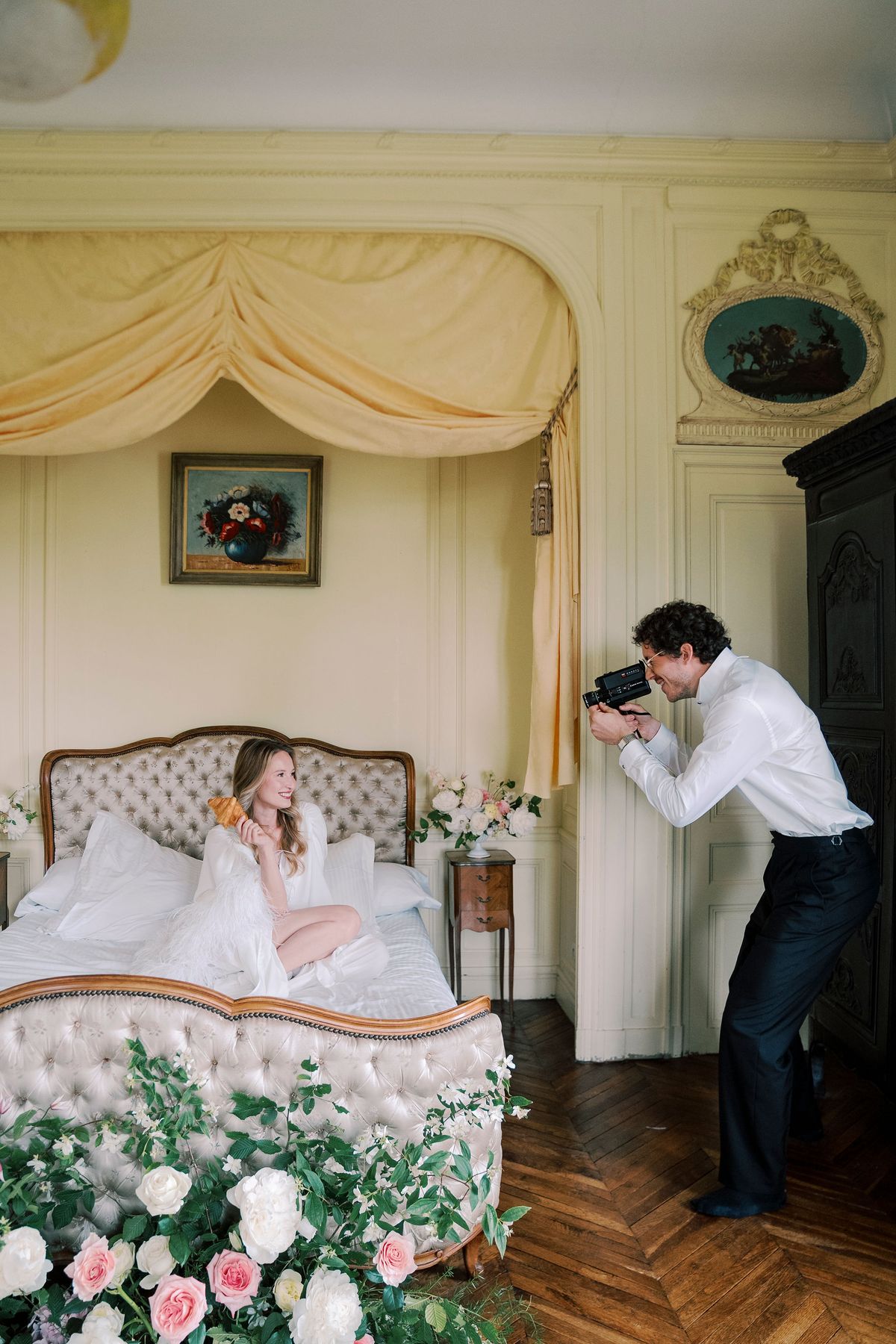 Lucien photographing Amélie on the canopied bed with a camera, roses in the foreground