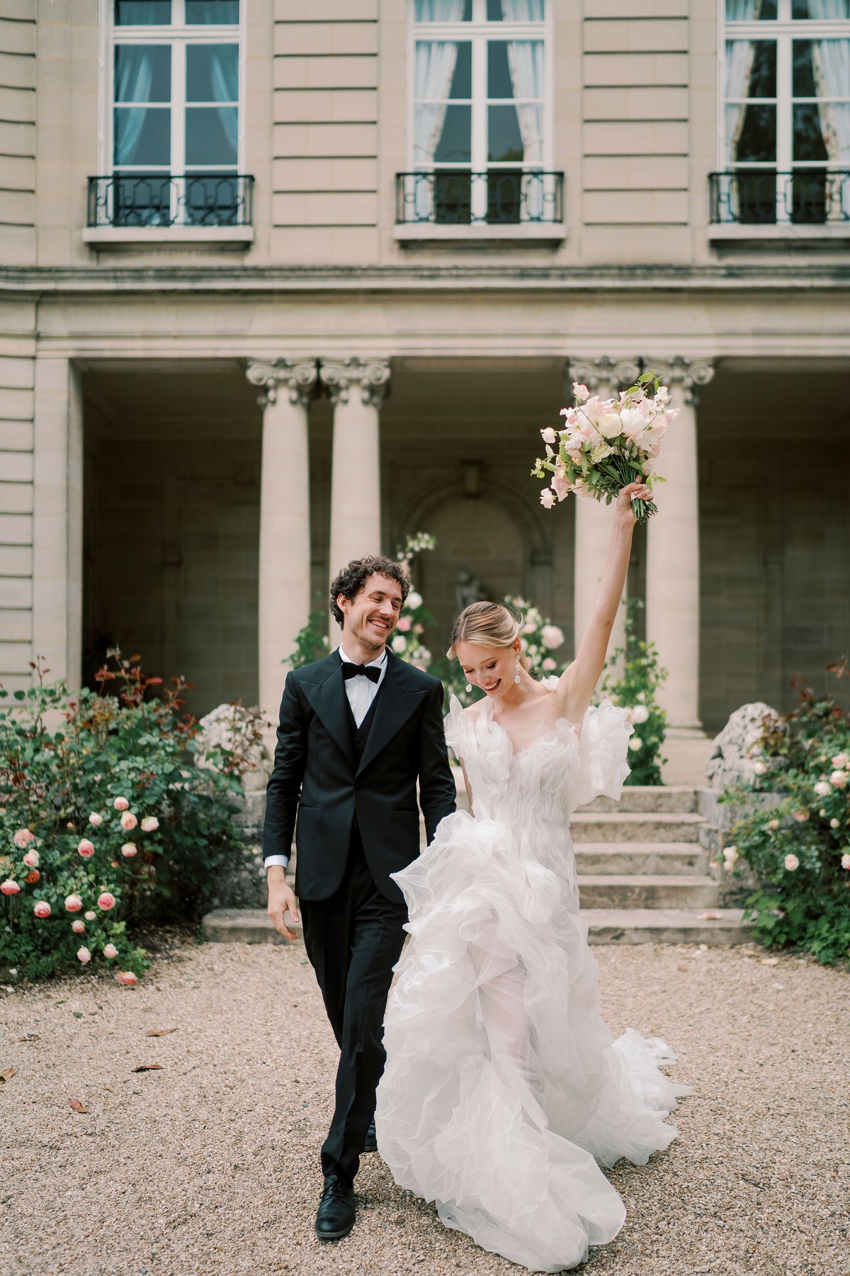 Amélie raising her bouquet as she and Lucien walk across the gravel from the peristyle