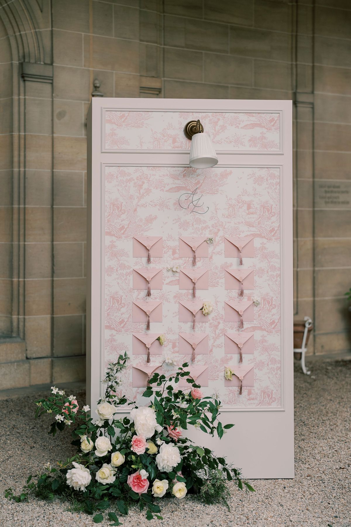 Seating chart panel in blush toile de Jouy with rows of pink envelopes and a wall sconce