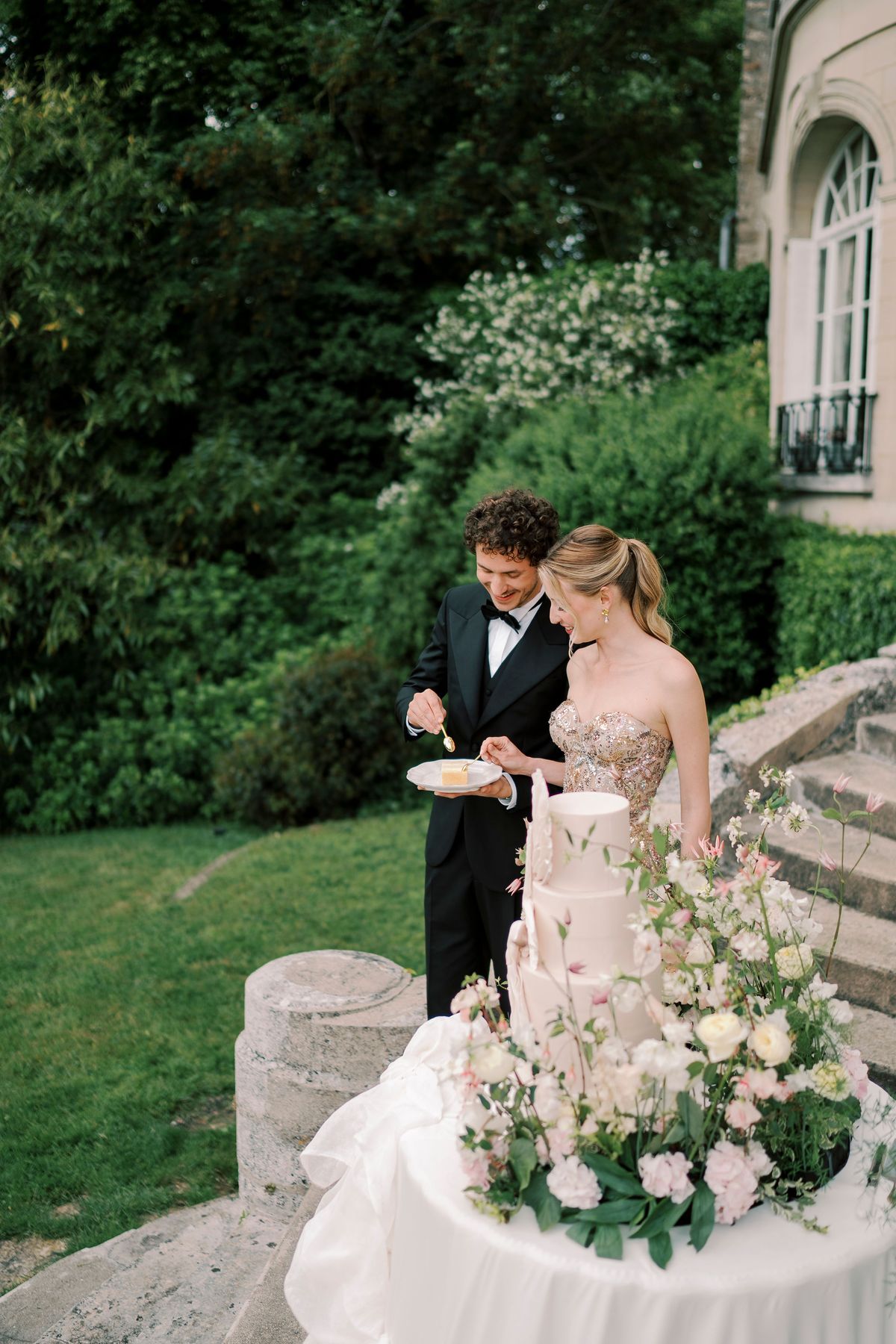 Amélie and Lucien cutting the tiered cake on the steps beside the lawn