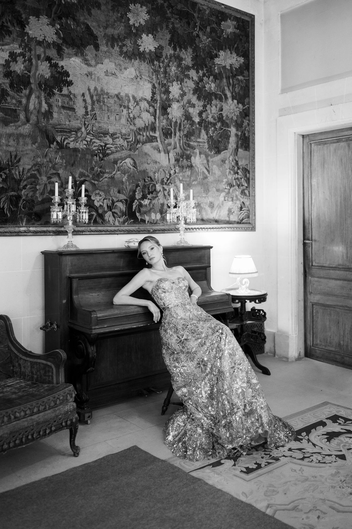Black and white portrait of Amélie in a sequined gown leaning on a piano beneath a tapestry