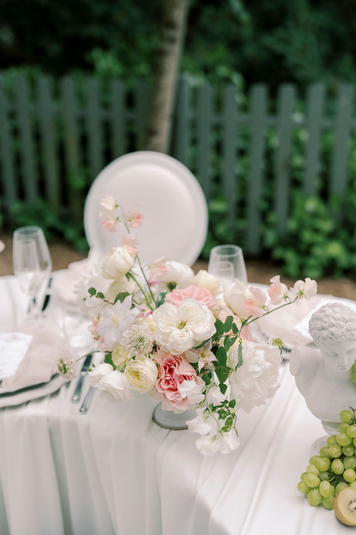 Pink and white roses beside glassware, a plaster bust and green grapes on the table