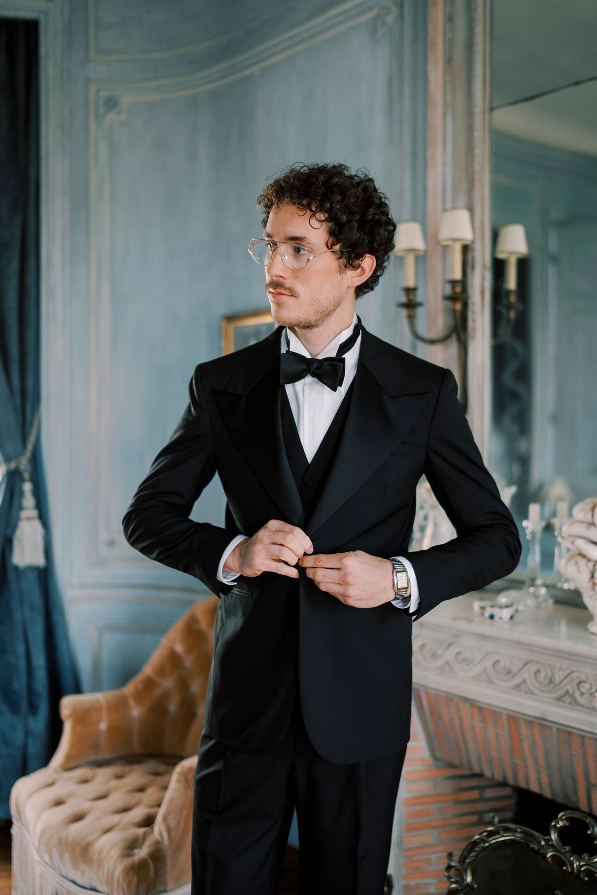 Lucien fastening his black tie jacket in a pale blue panelled room at Château du Prieuré