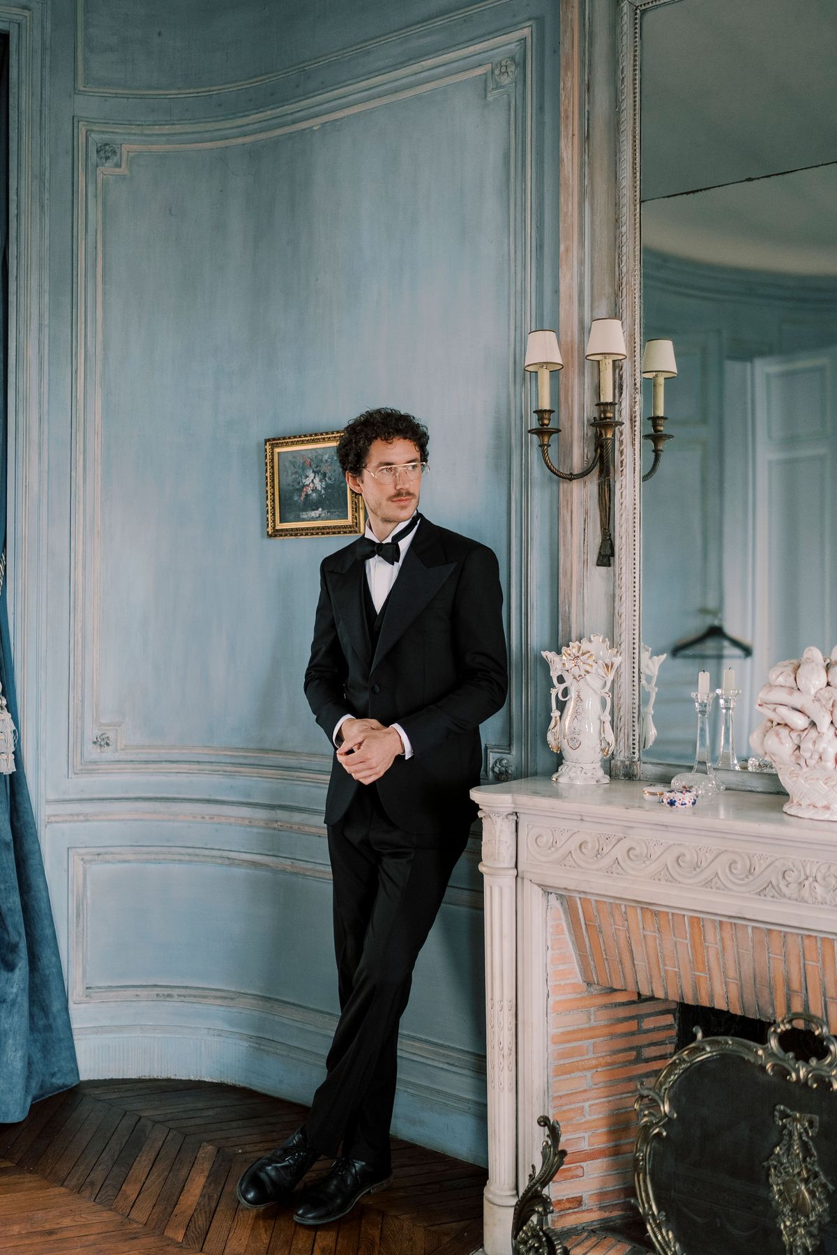 Lucien in black tie leaning on a marble fireplace in a pale blue panelled room