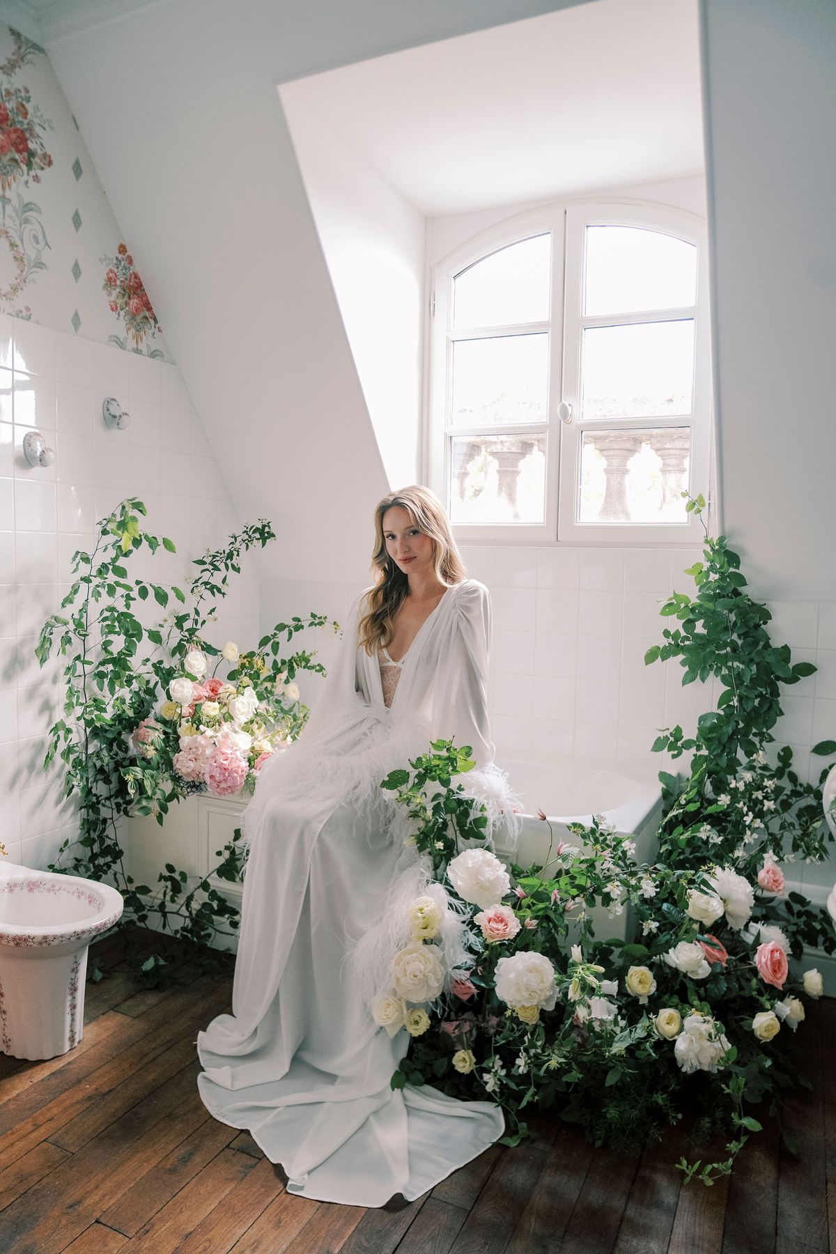 Amélie in a white robe beside the bathtub, surrounded by roses and climbing greenery