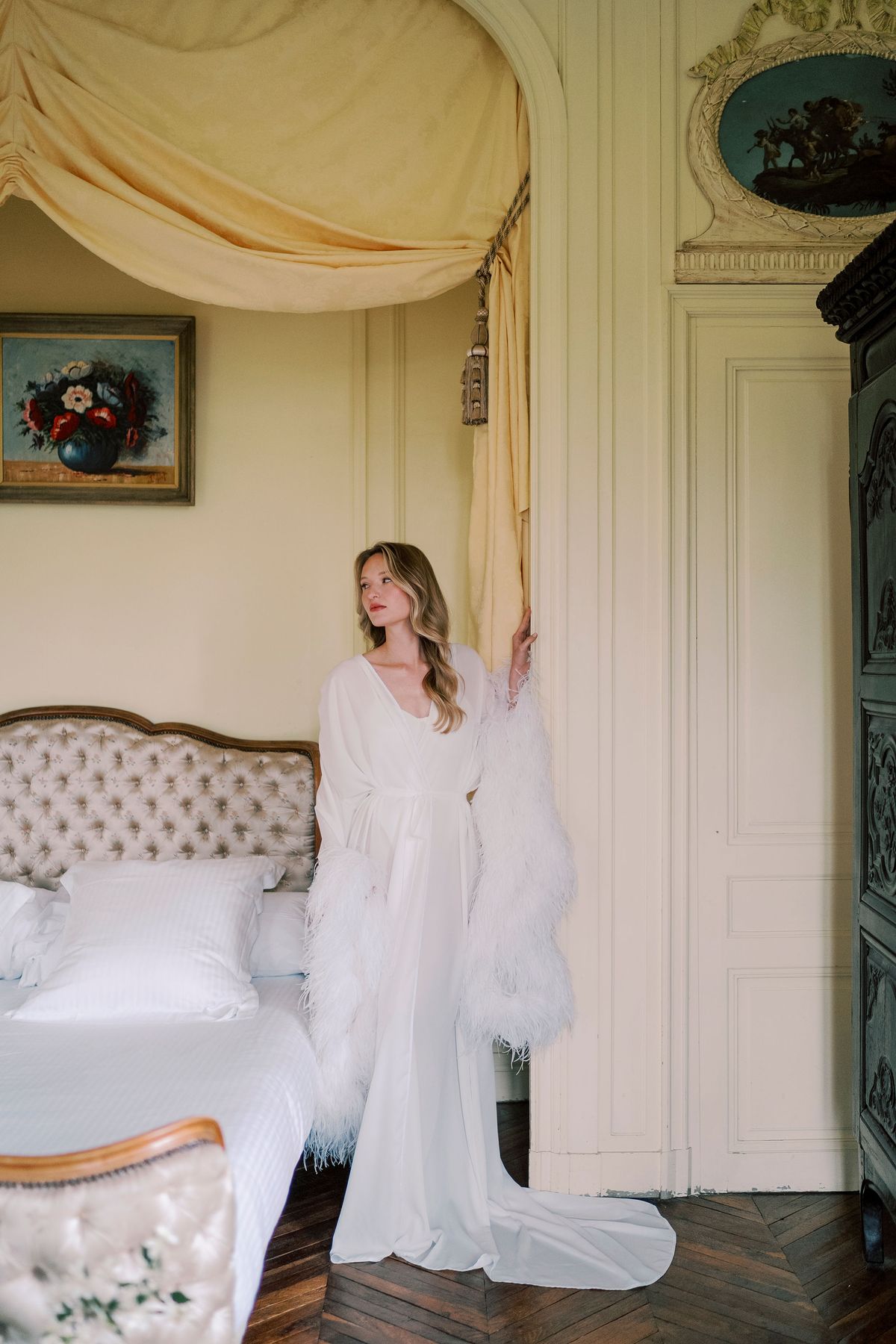 Amélie in a long white feathered robe leaning in the doorway of the yellow bedroom