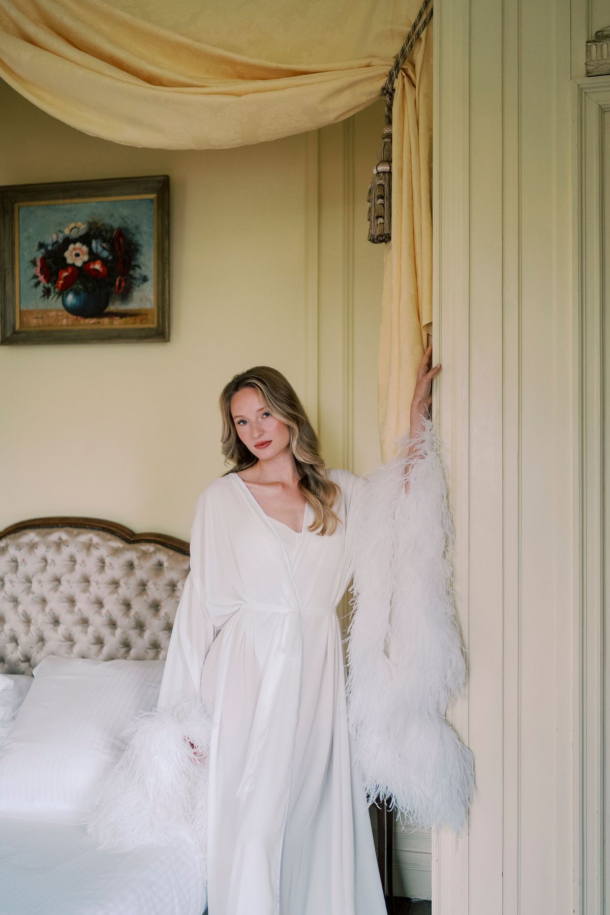Amélie in a white robe with feathered sleeves beside the canopied bed
