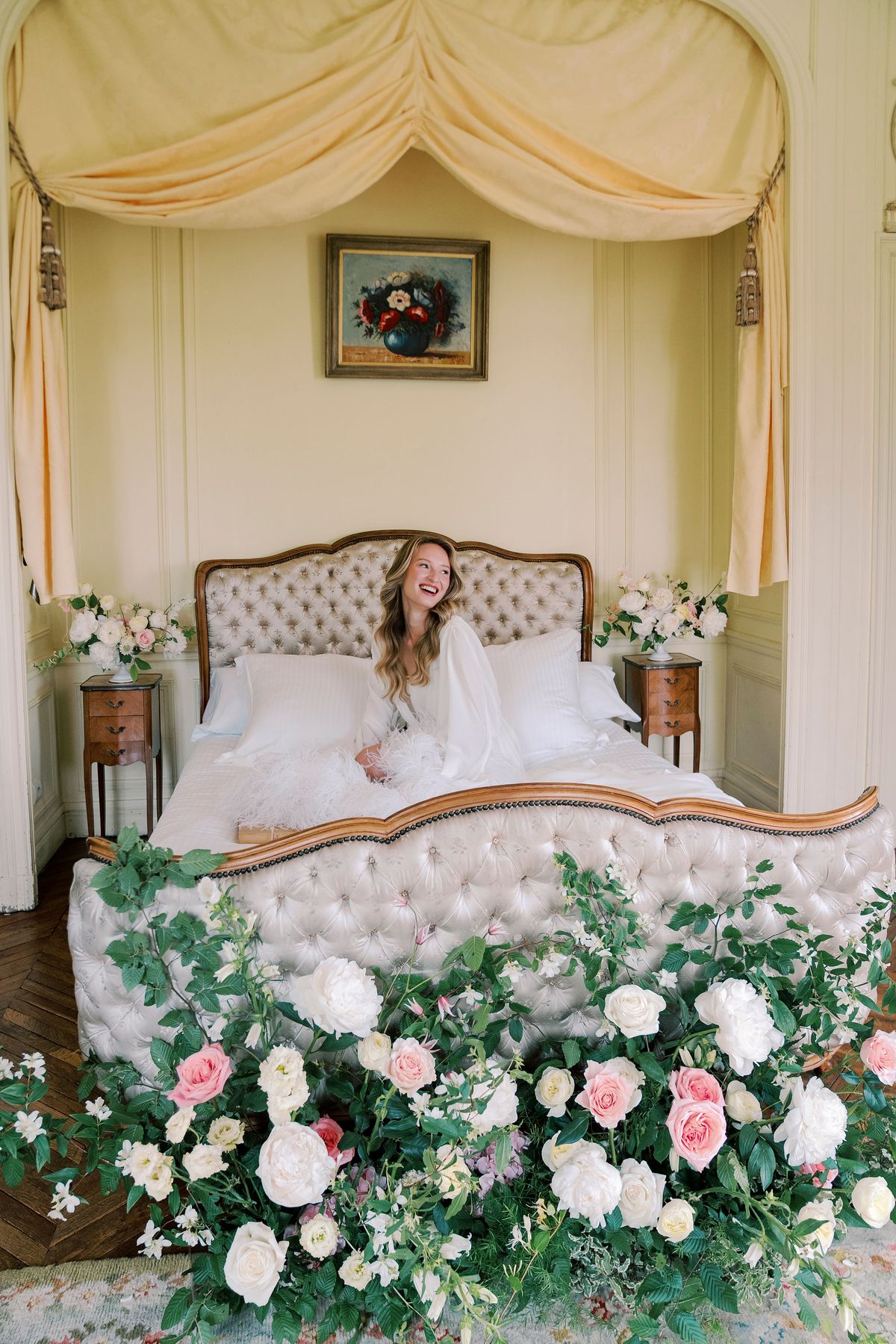Amélie laughing on the tufted canopied bed, garden roses and greenery at its foot