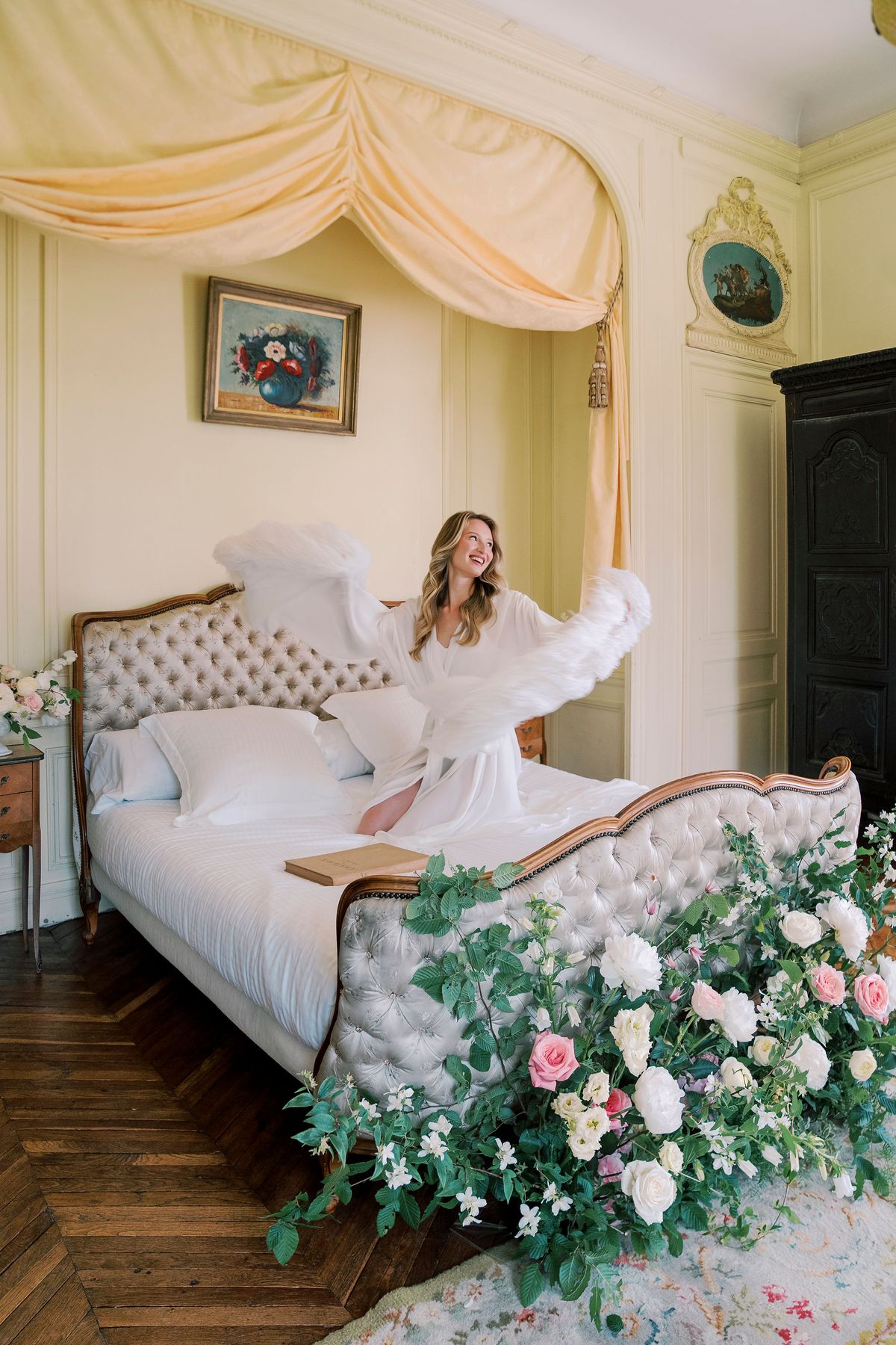 Amélie on the canopied bed, lifting the feathered sleeves of her white robe