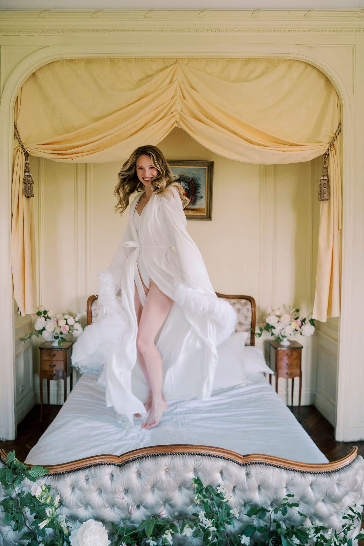 Amélie standing on the bed in a white feathered robe, roses along the foot of the bed