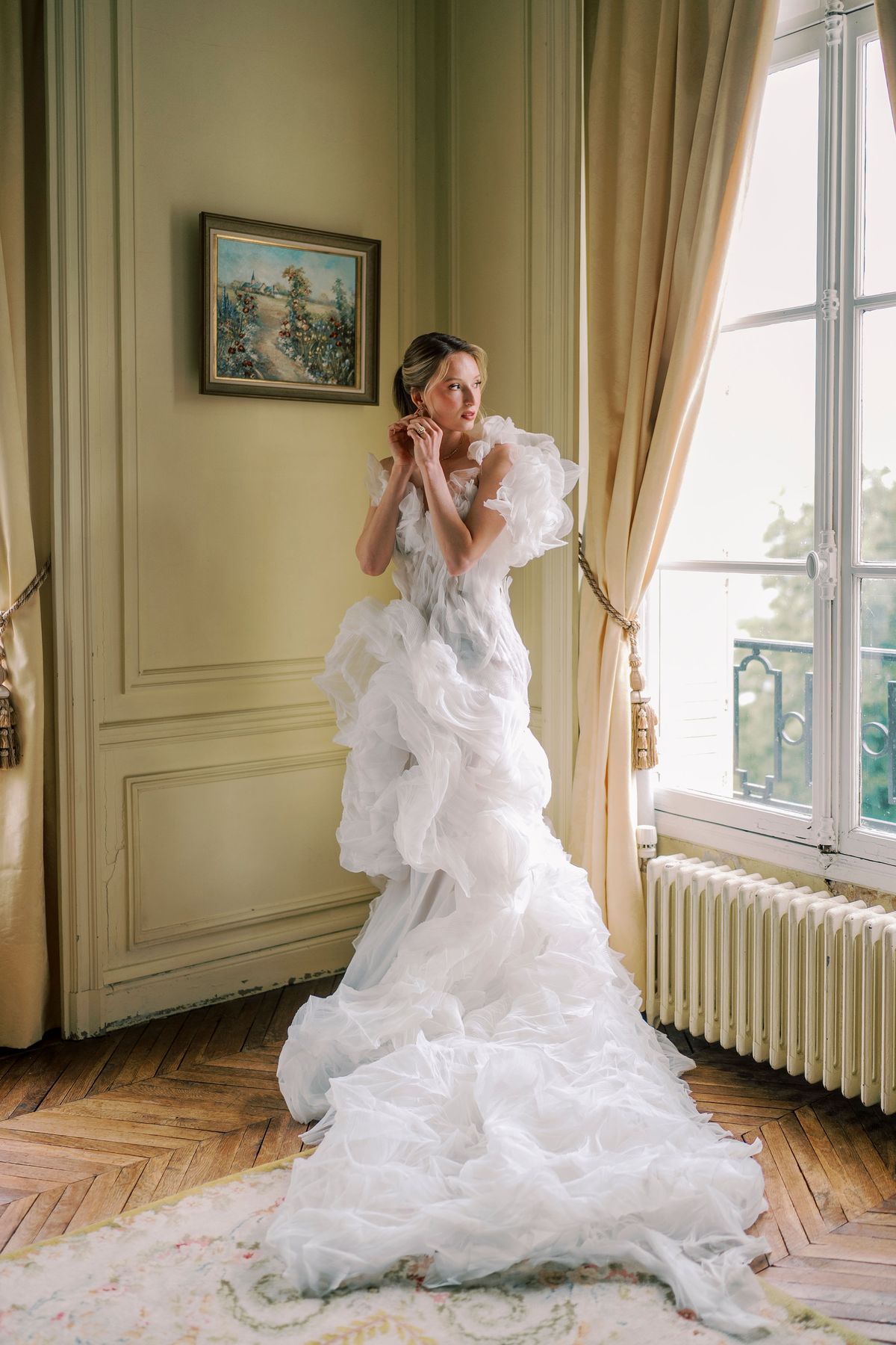 Amélie fastening an earring by a tall window in a yellow bedroom at Château du Prieuré, wearing her organza gown