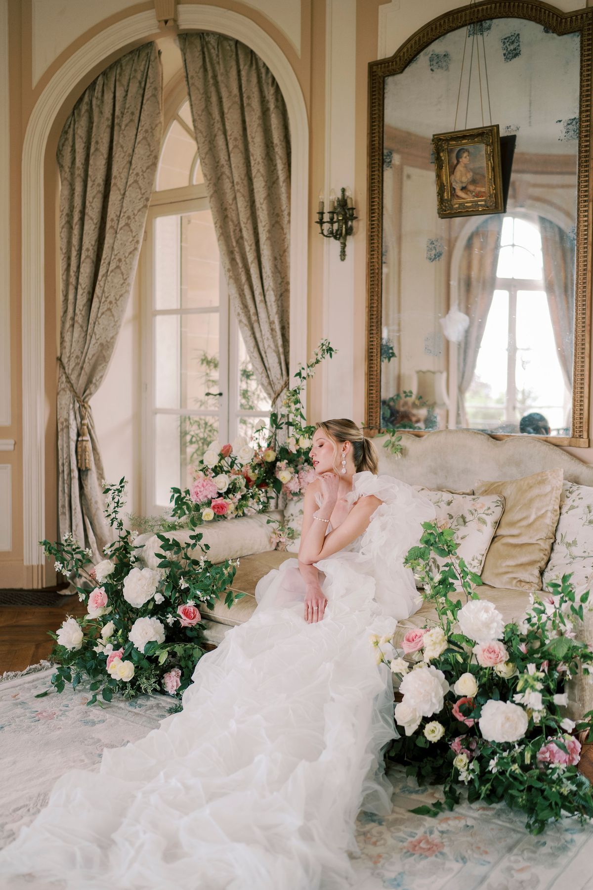 Amélie seated among floor-level roses in a salon, her organza train across the rug