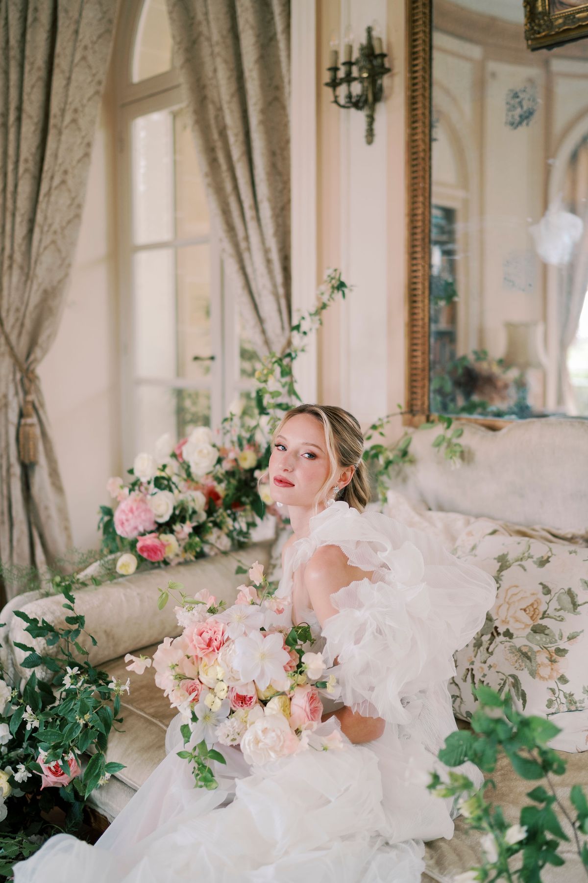 Amélie holding her bouquet on a pale sofa in a salon, roses around her