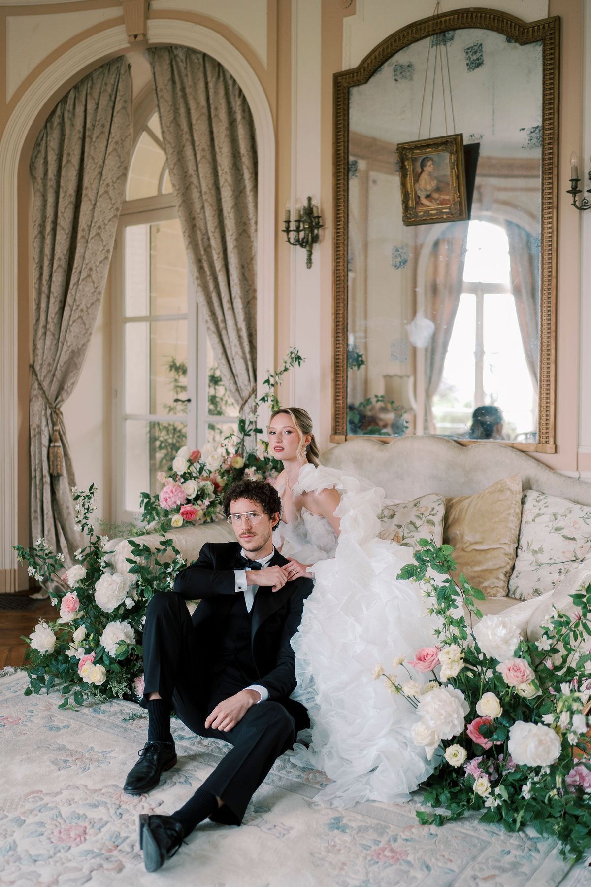 Lucien seated on the floor beside Amélie on a salon sofa, surrounded by roses