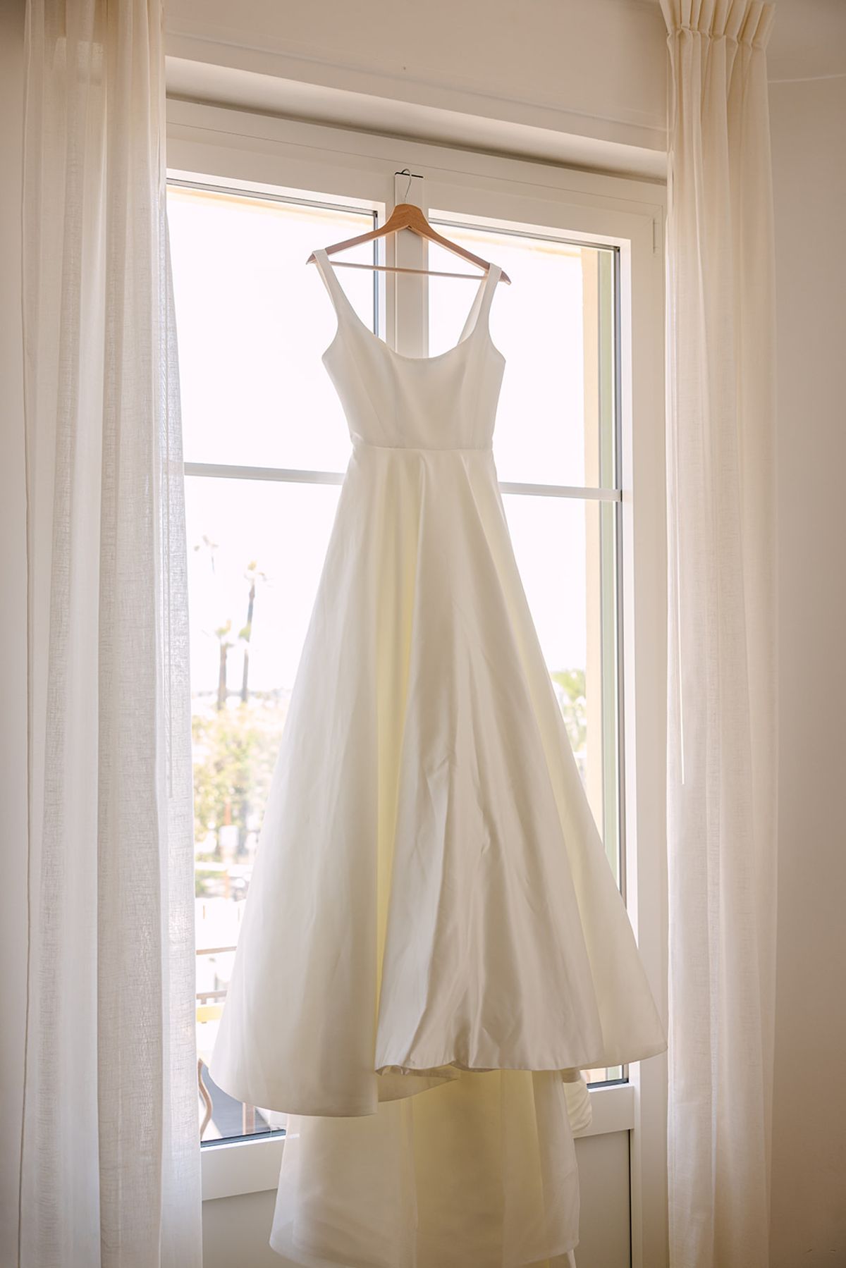 A white wedding dress with wide straps hangs from a window frame between sheer white curtains.