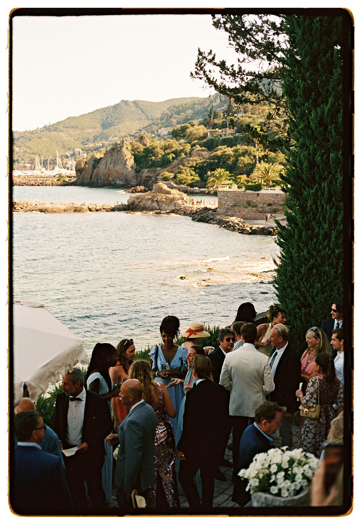 Seen from above, a crowd of dressed-up guests gathers on a terrace beside the water, with a rocky coastline and hills beyond.