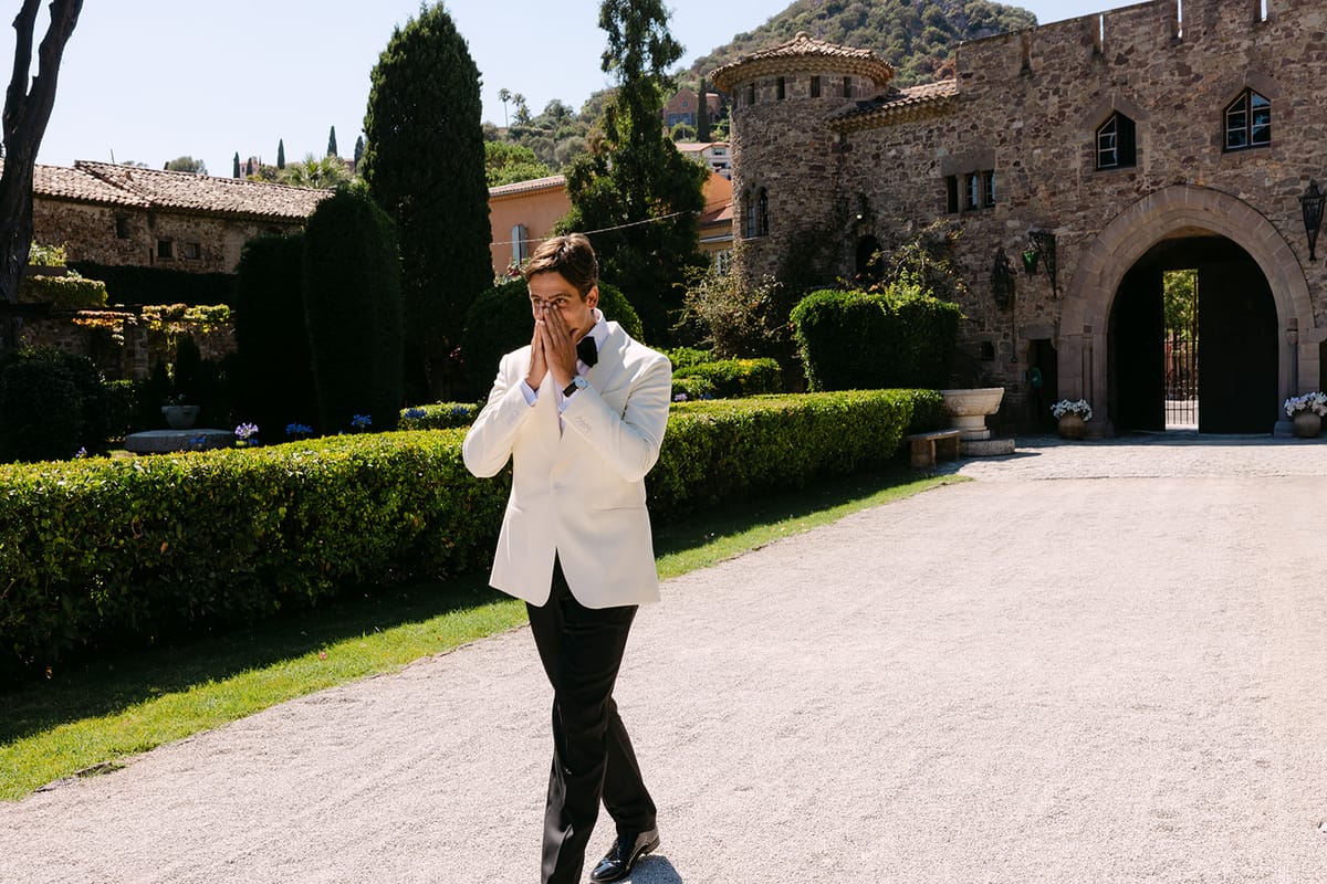 The groom in a white dinner jacket walks along a gravel drive with his hands to his face, a stone gateway behind.