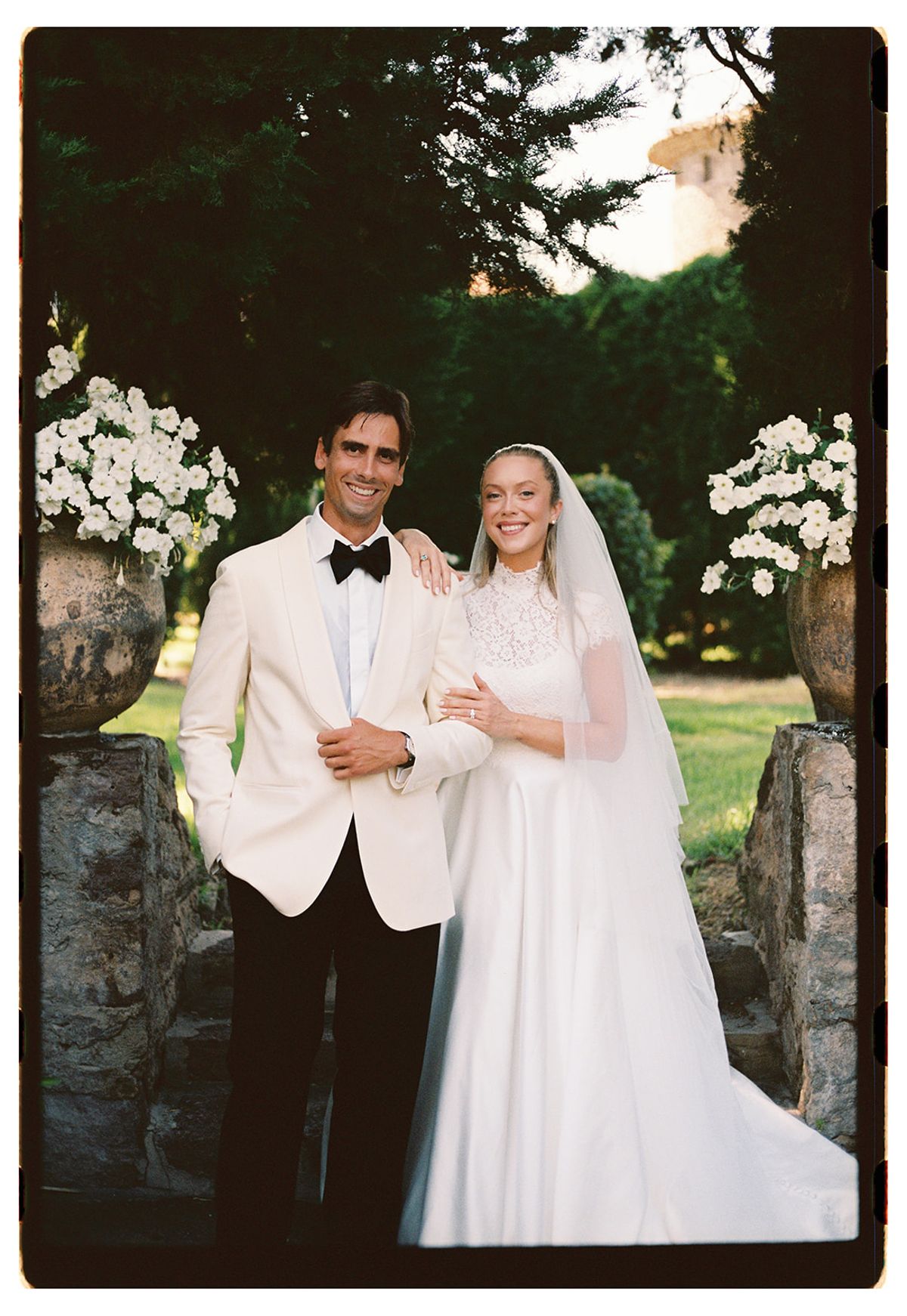 The groom in a white dinner jacket and the bride in a long veil pose together between urns of white flowers in a garden.