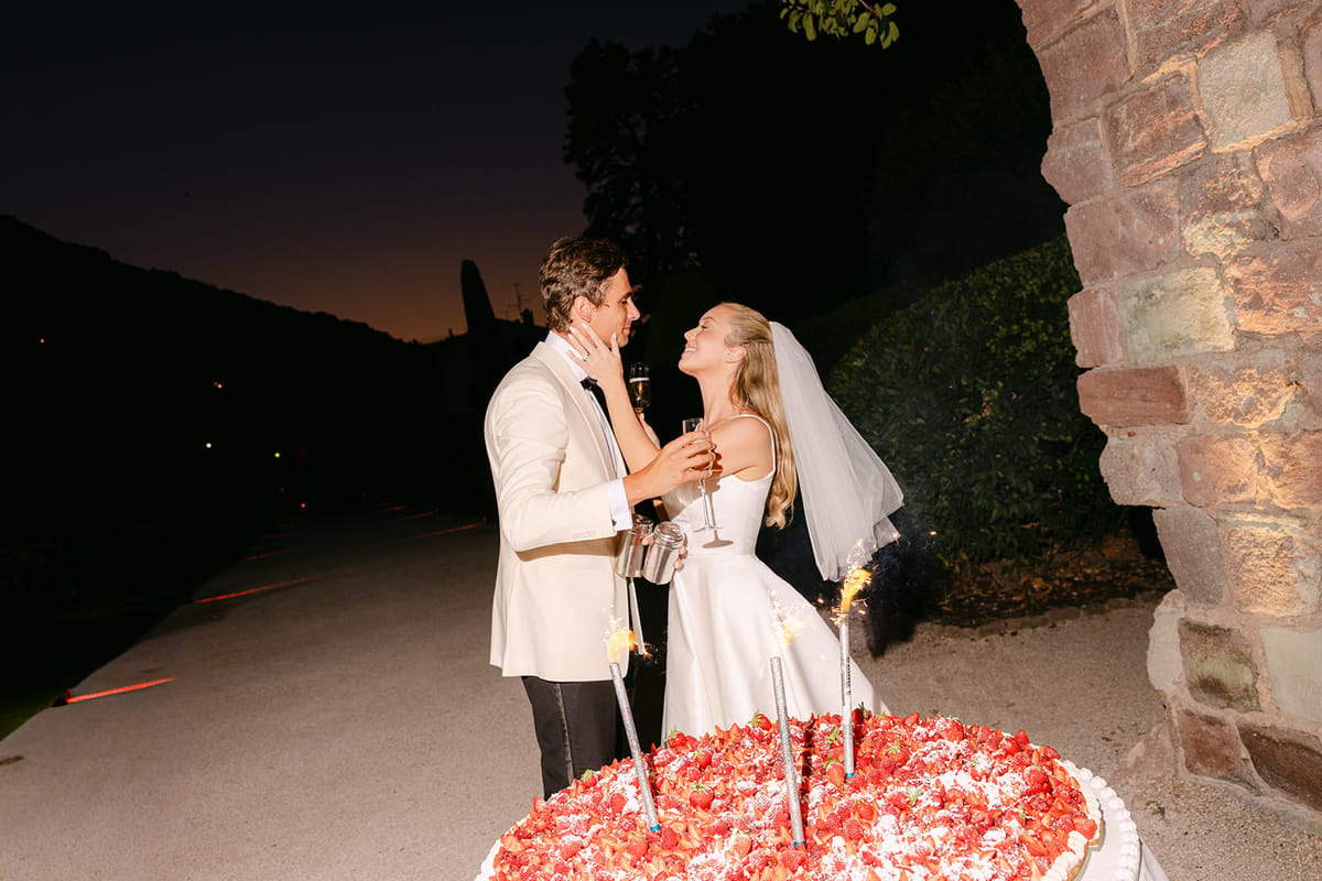 The couple stand close together behind a strawberry cake with sparklers, outdoors at night beside a stone arch.