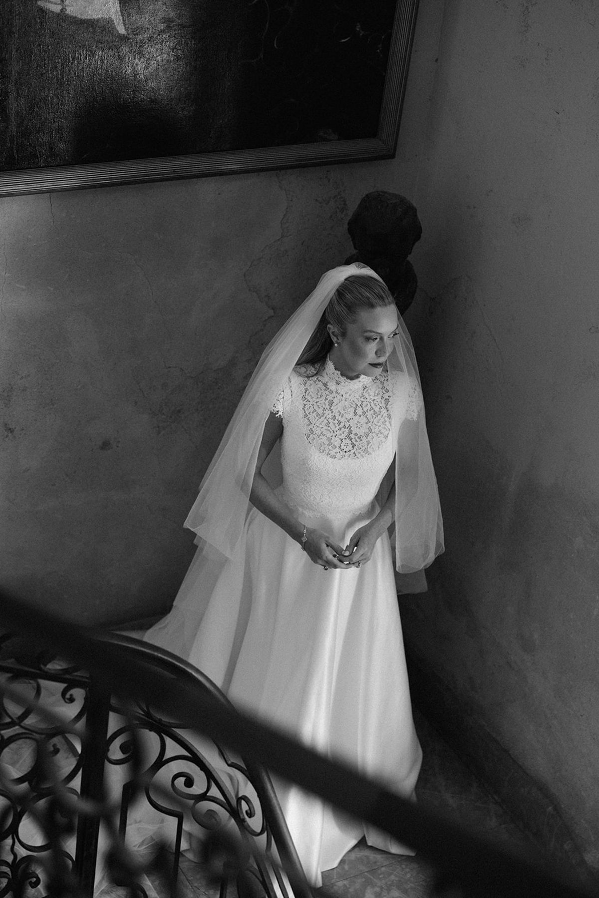 In black and white, the bride in a veil and lace-topped gown stands on a stone staircase beside an ornate iron railing.
