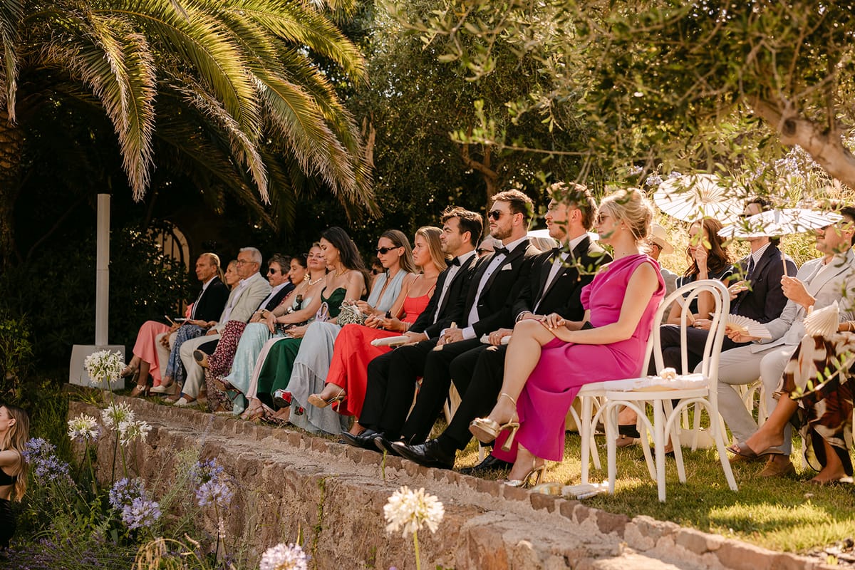 Guests in colourful outfits and sunglasses sit on white chairs in a sunlit garden beneath palm trees, watching the ceremony.
